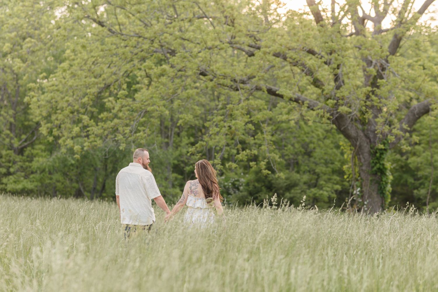 Two people hold hands while walking through a tall grass meadow surrounded by green trees on a summer evening.