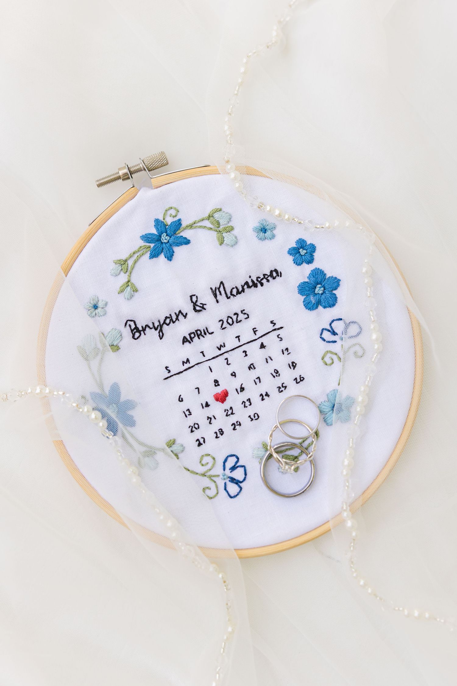 Embroidered wedding hoop art featuring a calendar, blue flowers, and wedding rings on white fabric with delicate details.