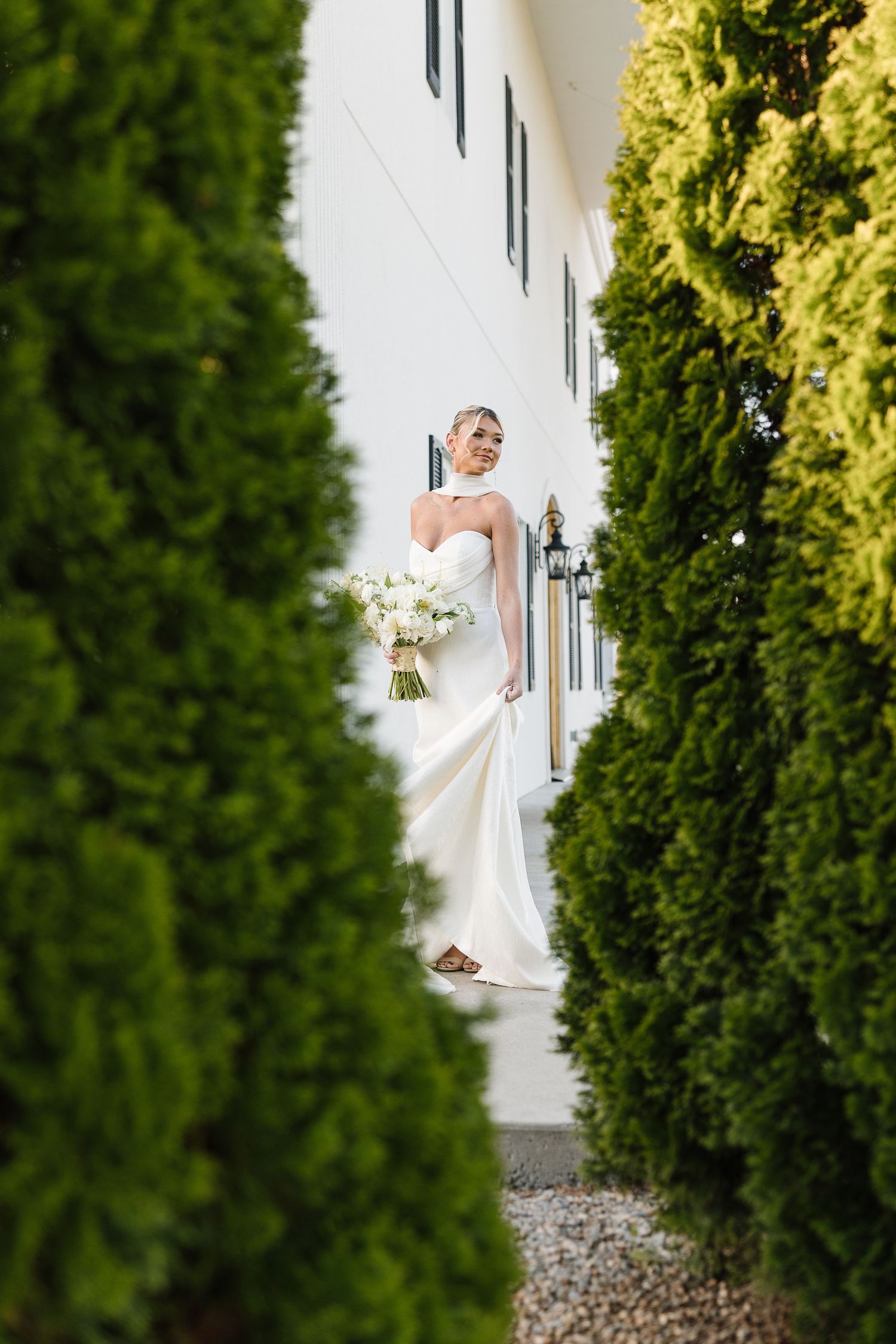 A bride in a flowing white gown poses elegantly between tall evergreen bushes outside a white building.