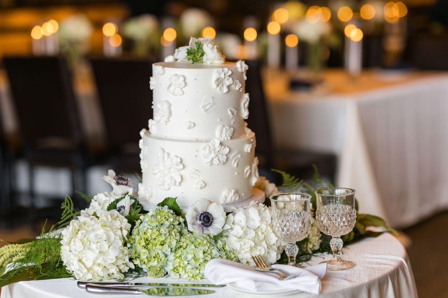 A white wedding cake with floral decorations sits on a table with bokeh lights in the background.