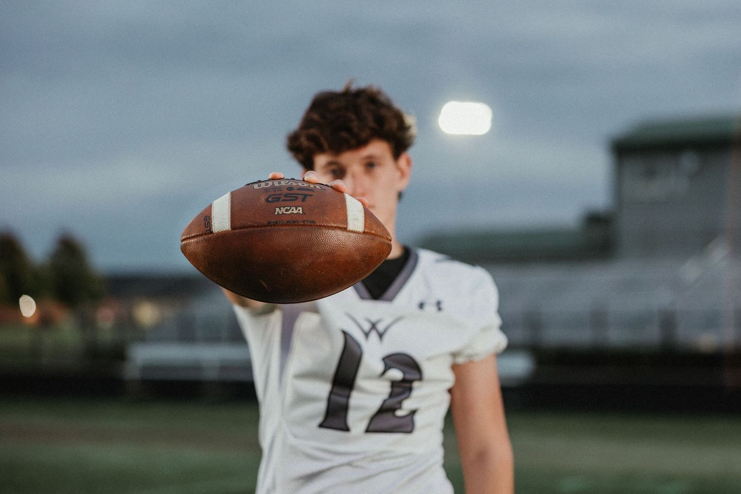 Football player in white jersey tosses ball in dramatic sunset lighting at stadium.