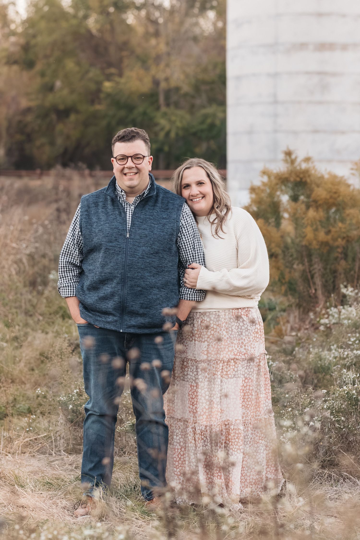 A couple poses outdoors in casual fall attire with a cream sweater and navy vest against an autumn backdrop.