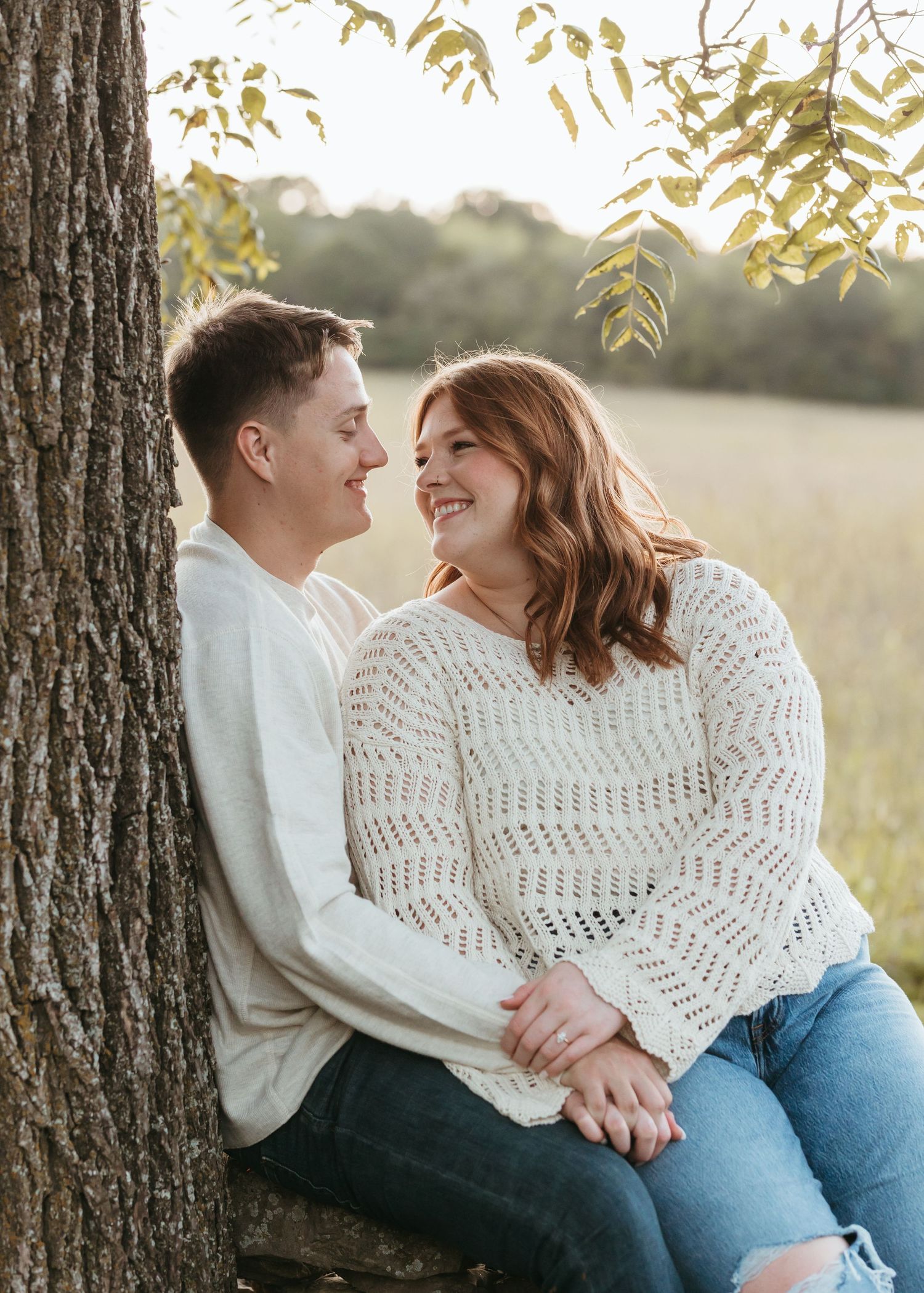 A couple shares an intimate moment while sitting by a tree in warm sunlight.
