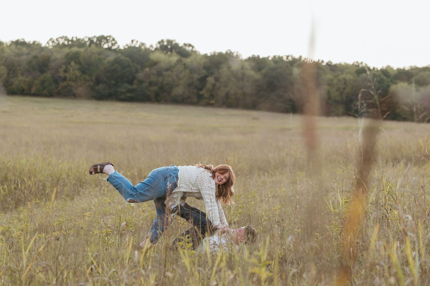 A couple playfully poses in a golden field with tall grass and trees in the background.