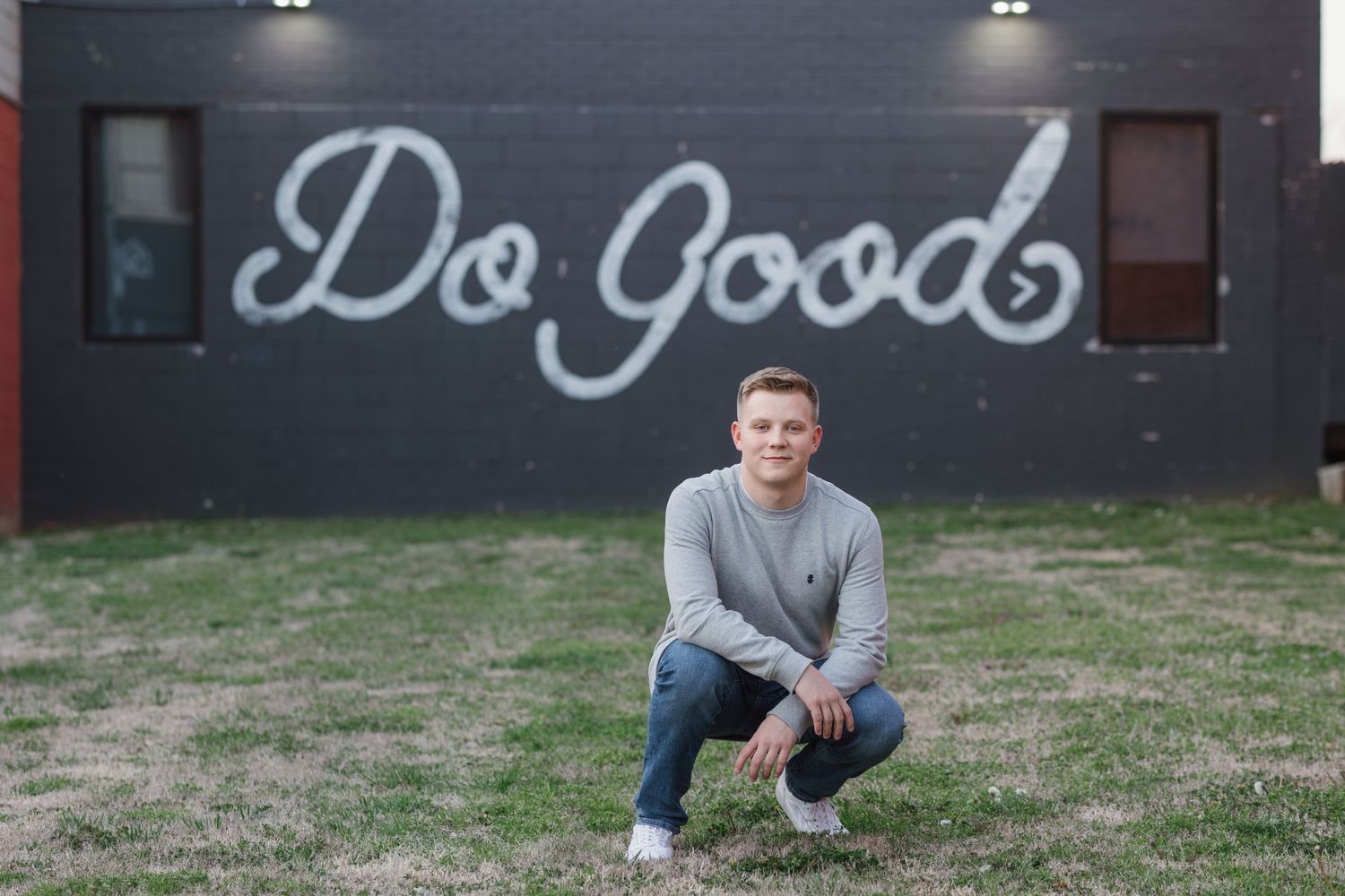 Person in gray sweater crouching in front of wall with 'Do Good' neon sign.