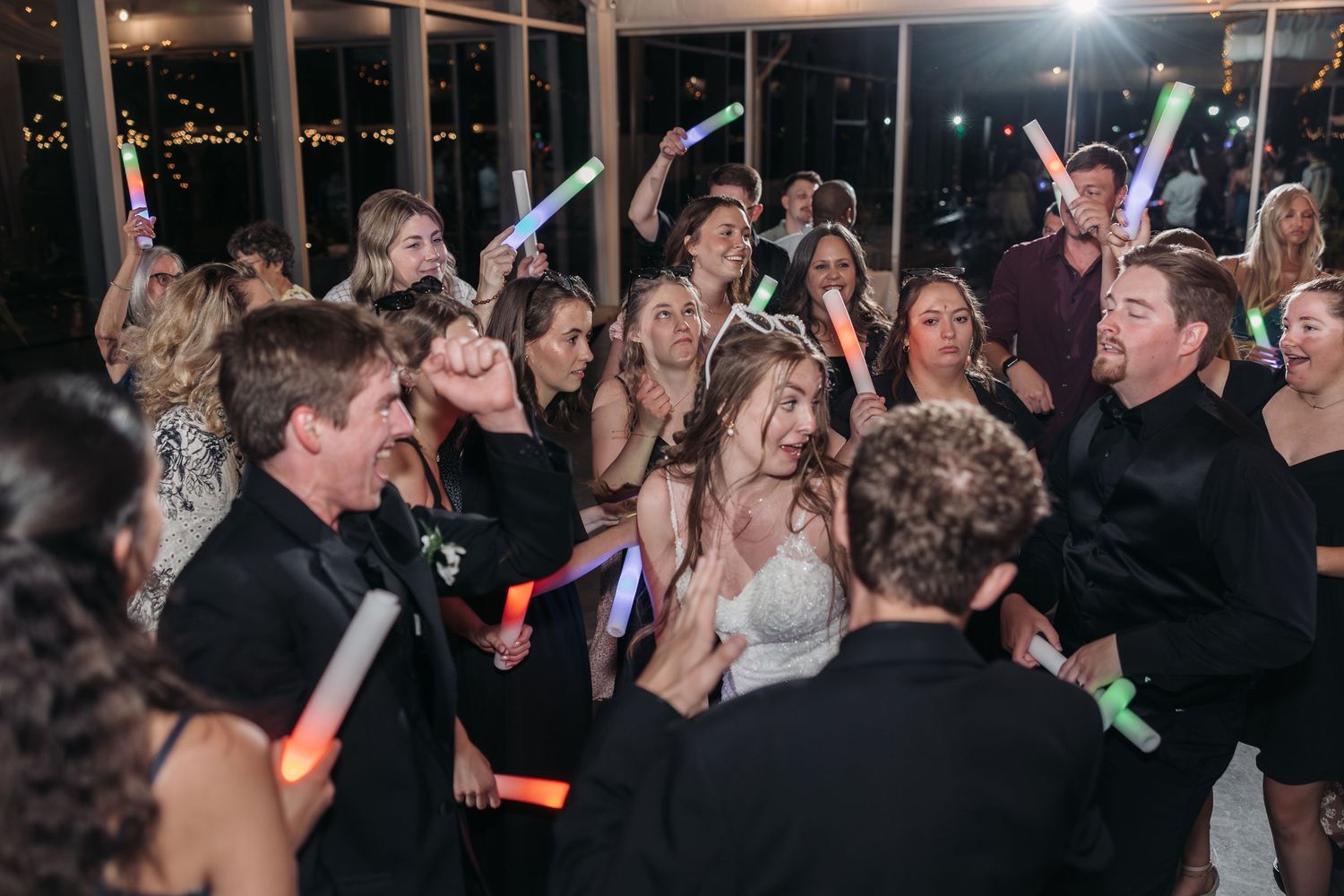 A large group of wedding guests dancing and celebrating with colorful light sticks.