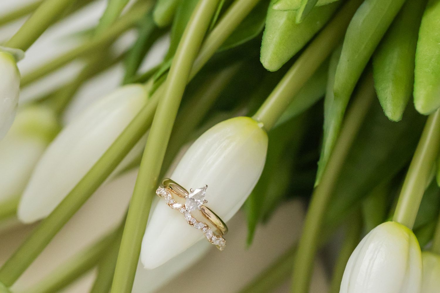 Gold wedding and engagement rings with diamond accents resting on white tulip bouquet.