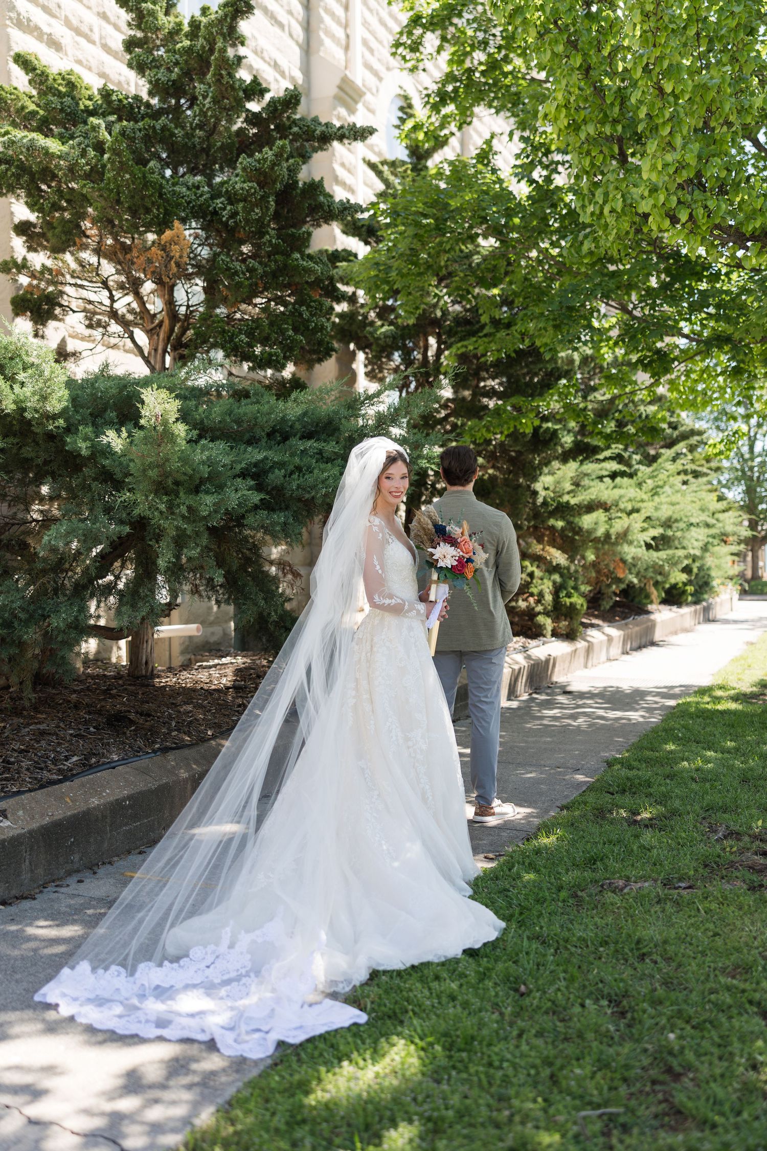 Bride in white gown and veil stands outdoors while groom walks ahead on a garden path near a building.