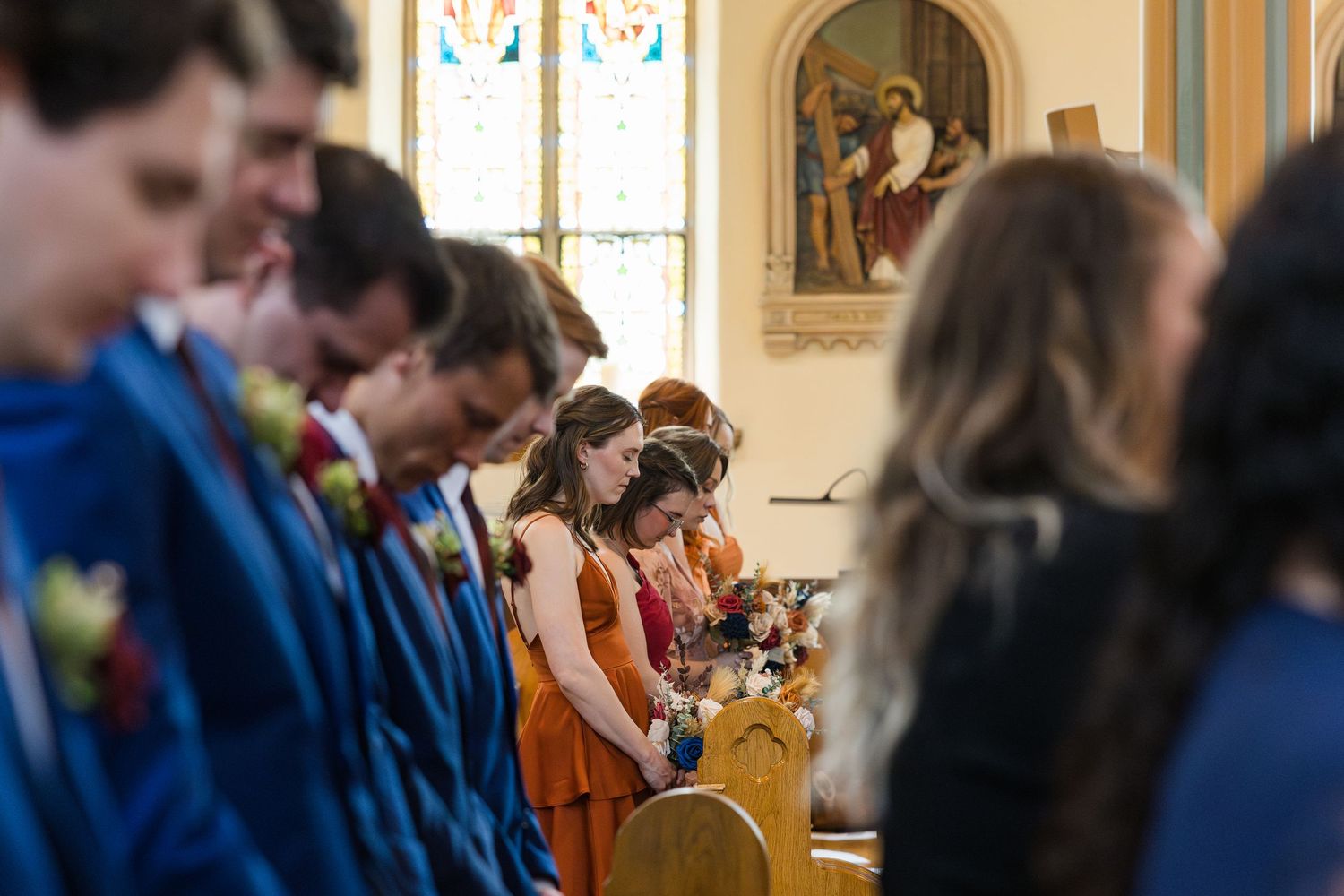 Wedding party members bow their heads in prayer during a church ceremony, dressed in blue suits and orange dresses.