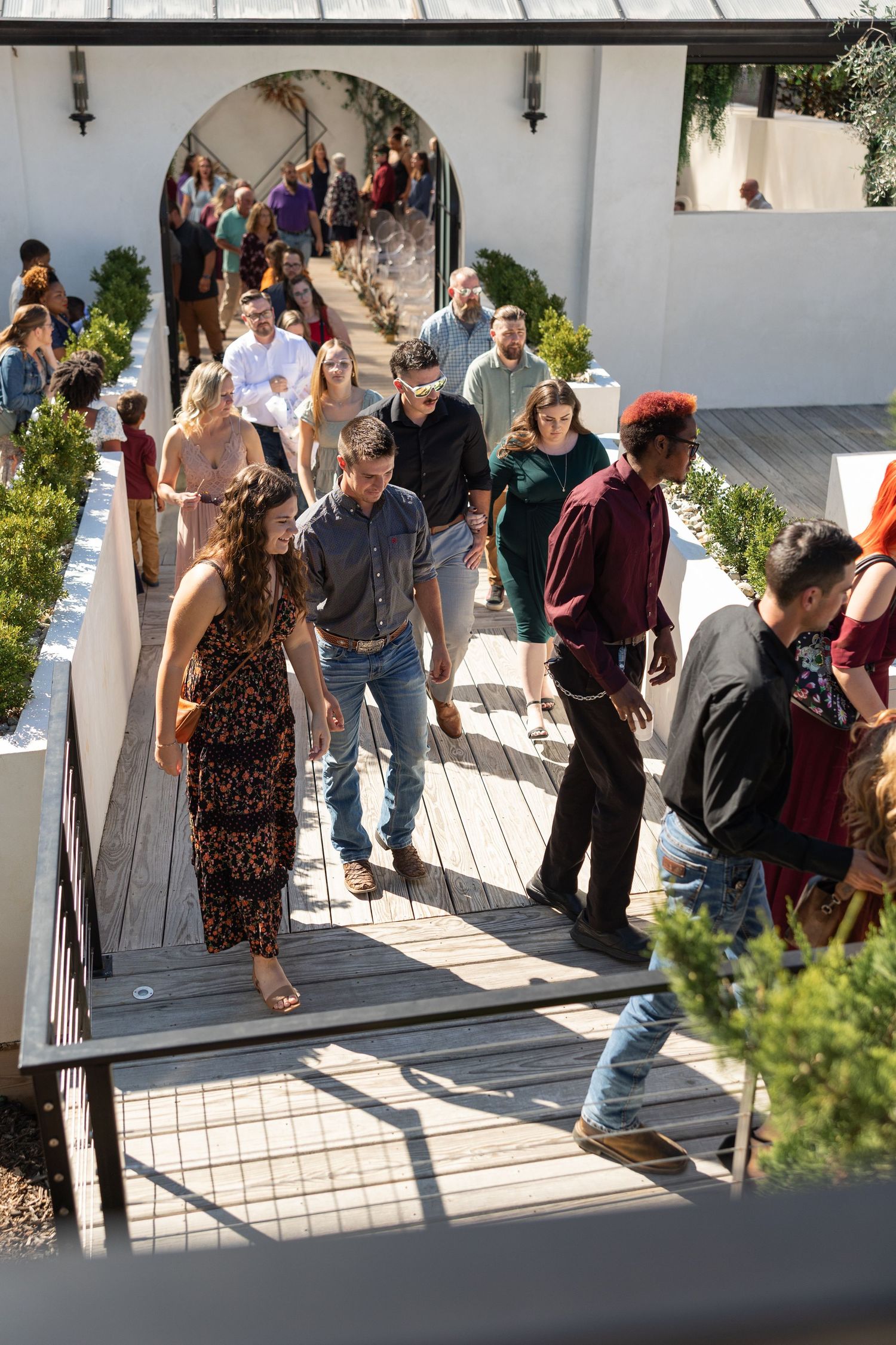 Wedding guests walk along a wooden pathway through a modern outdoor venue after the ceremony.