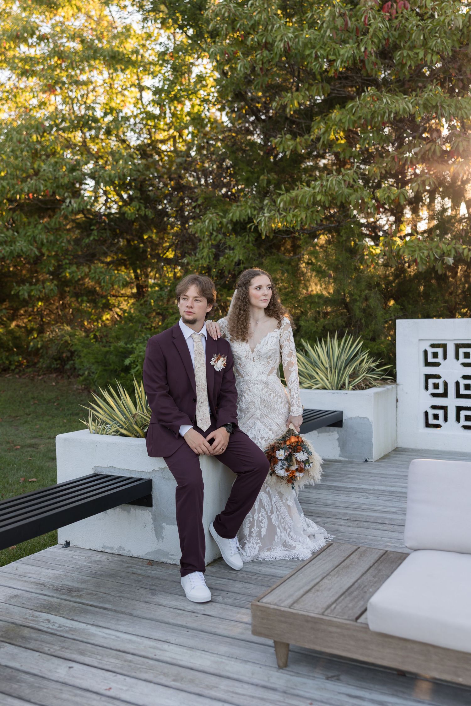 Bride and groom in burgundy suit sit together on outdoor deck at golden hour sunset.