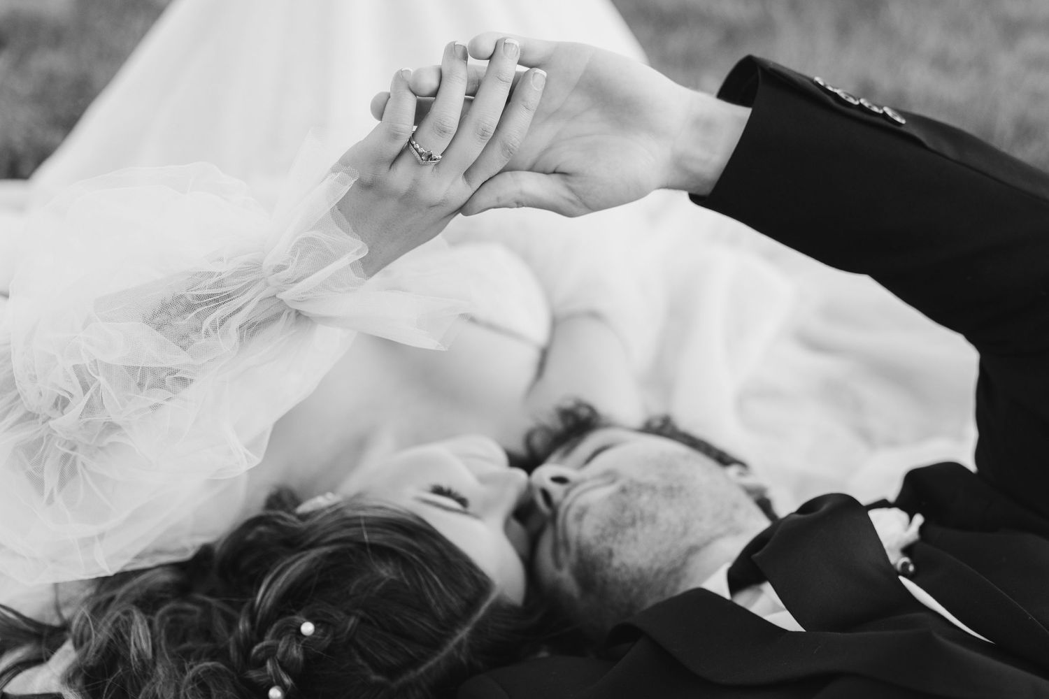 Black and white wedding photo of bride and groom lying in grass, holding hands with rings visible.