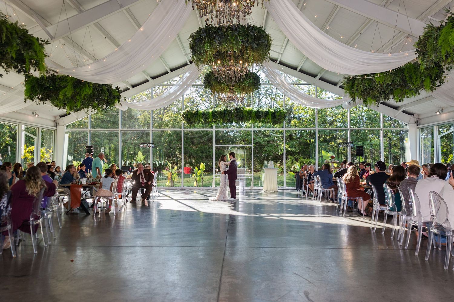 Elegant greenhouse wedding ceremony with chandelier, lush greenery, and guests seated in white chairs on a polished floor.