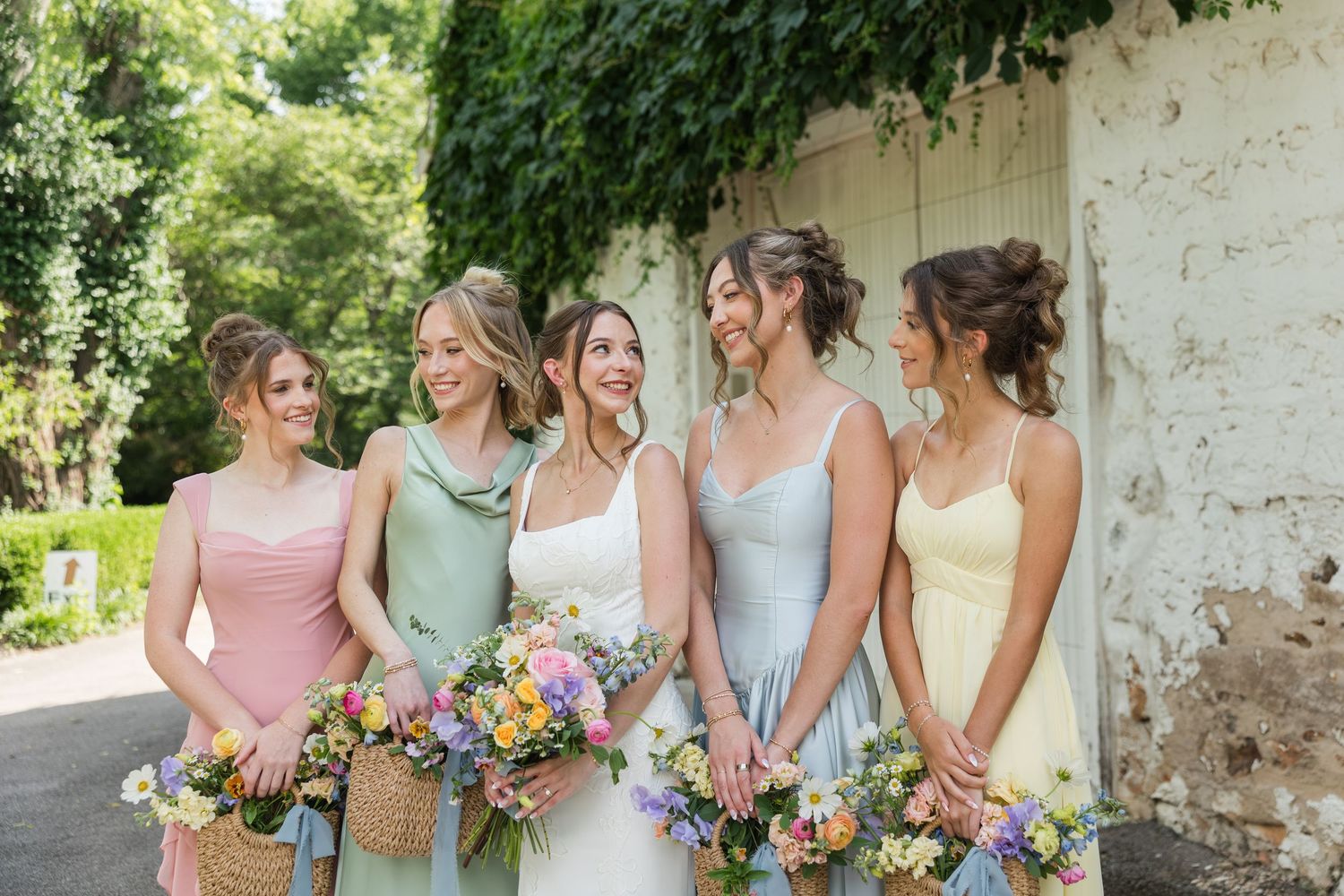 Bride and four bridesmaids in pastel dresses holding colorful wildflower bouquets and wicker baskets outdoors.