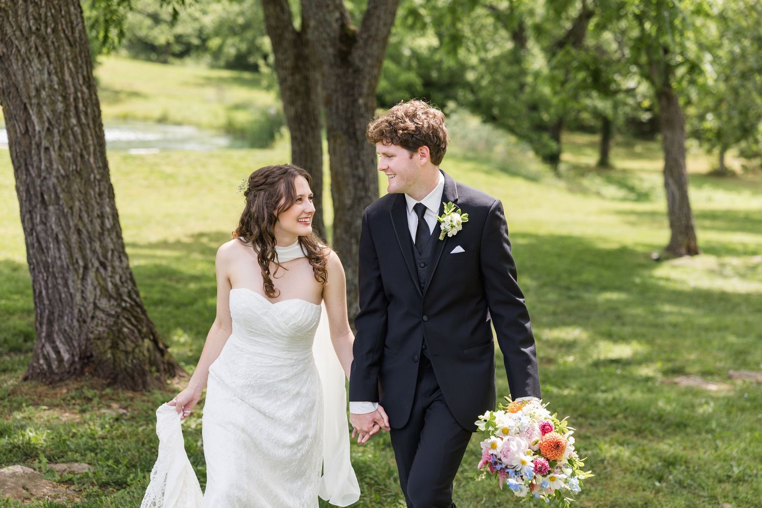 Bride and groom walking hand in hand outdoors among trees, bride holding colorful bouquet in white gown.