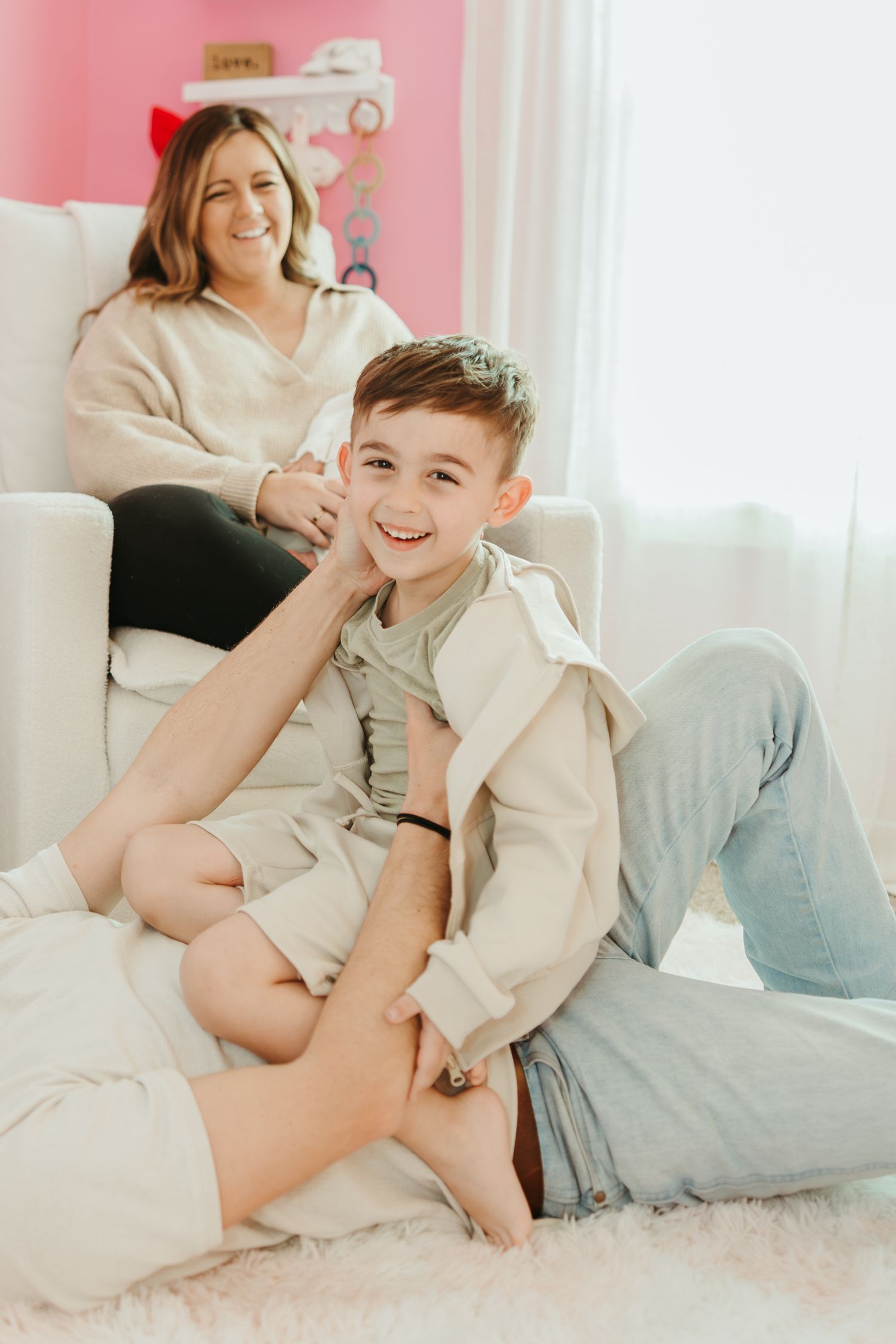 A candid moment of laughter and playfulness on a plush carpet against a pink wall with natural lighting.