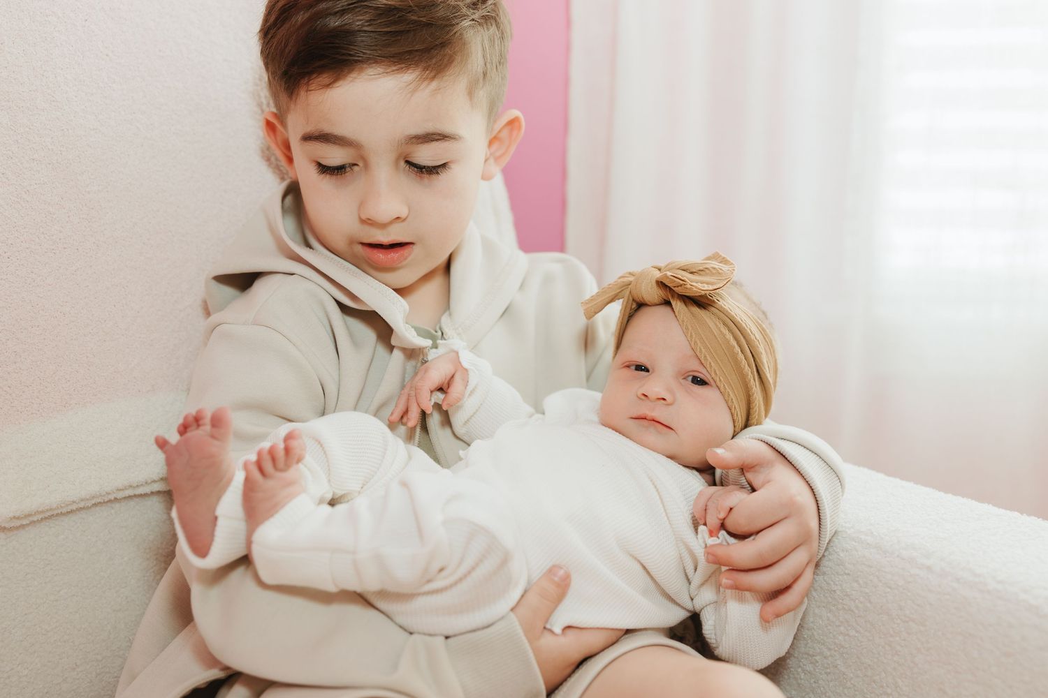 Two children sitting on white couch wearing cream colored knit sweaters in soft pastel interior.