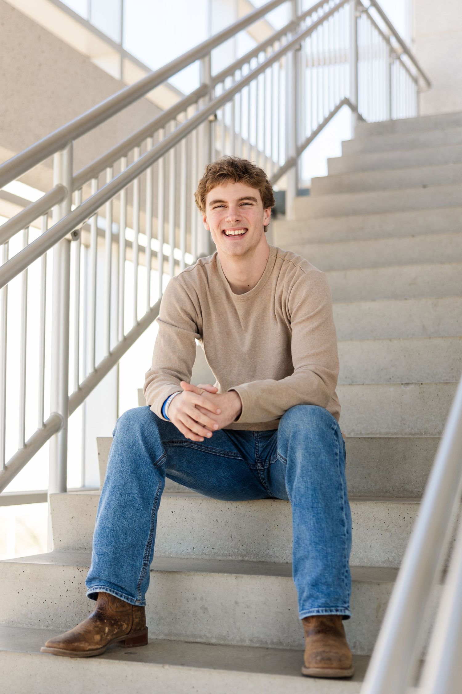 Student in beige sweater and jeans sits casually on concrete stairs by metal railing in bright interior space.