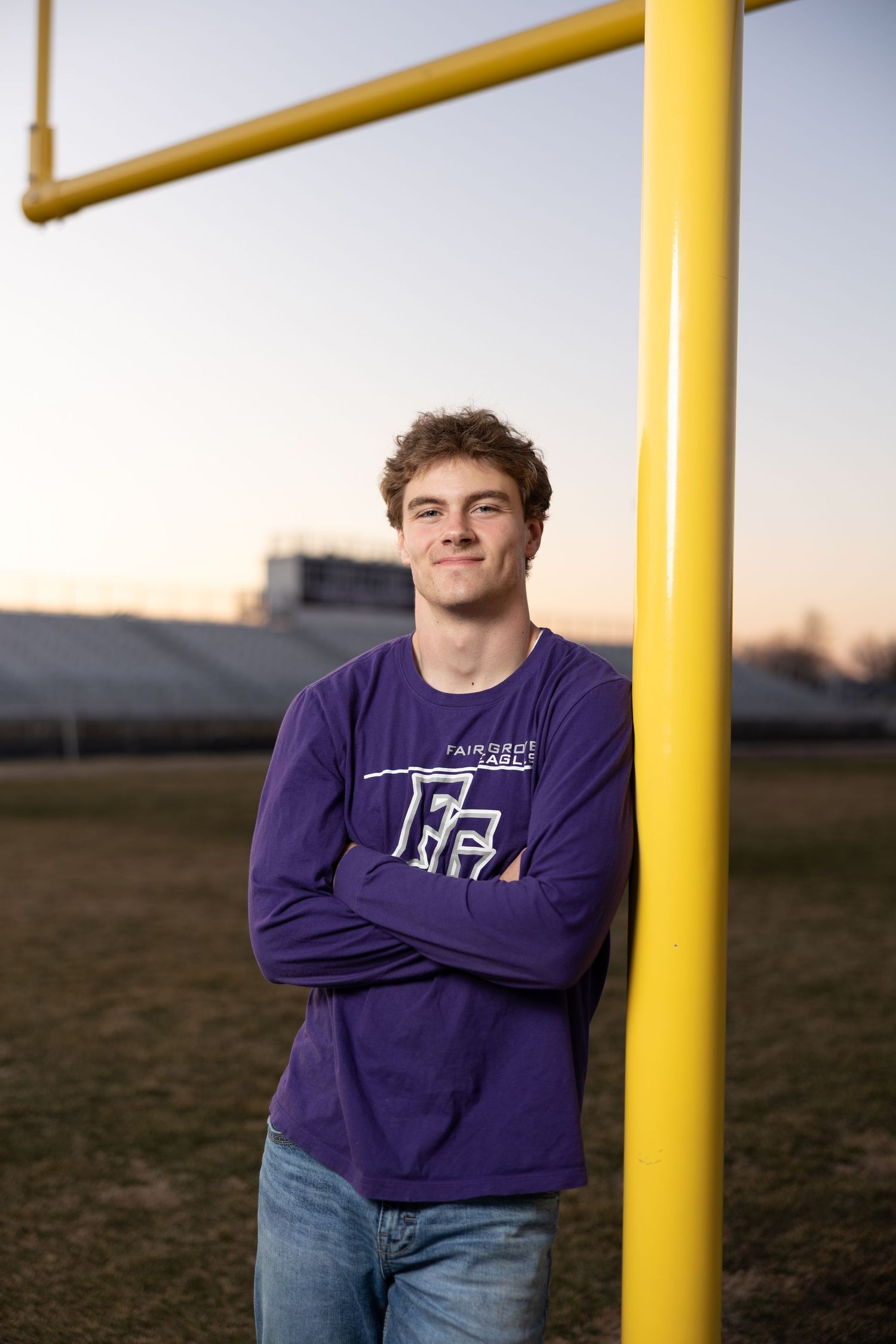 Student wearing purple long sleeve shirt and jeans stands next to yellow football goal post at sunset on sports field.