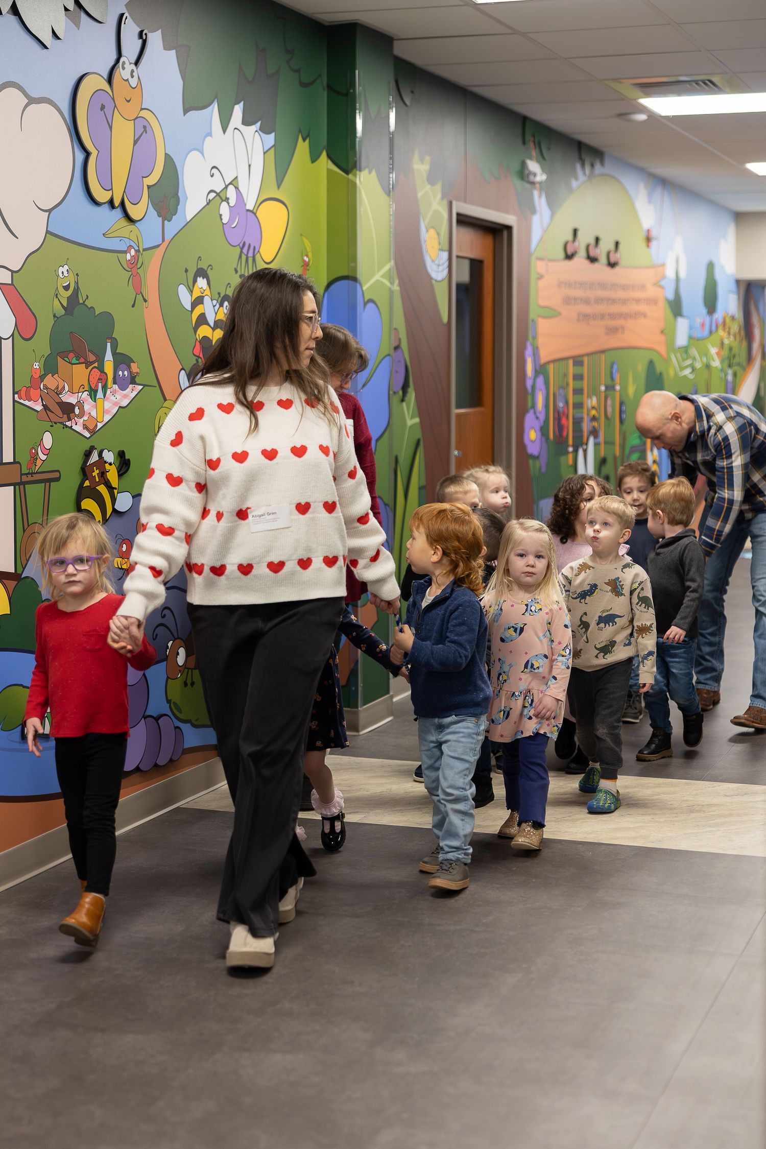 Children following teacher in heart-patterned sweater down whimsically decorated corridor.
