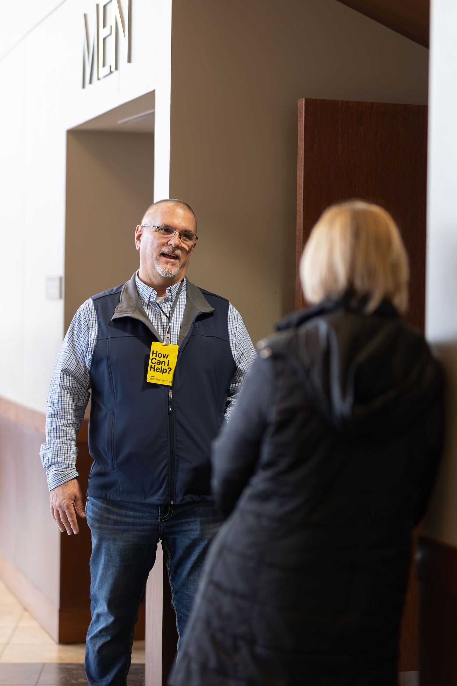 Person wearing event badge standing near doorway marked 'MEN'.