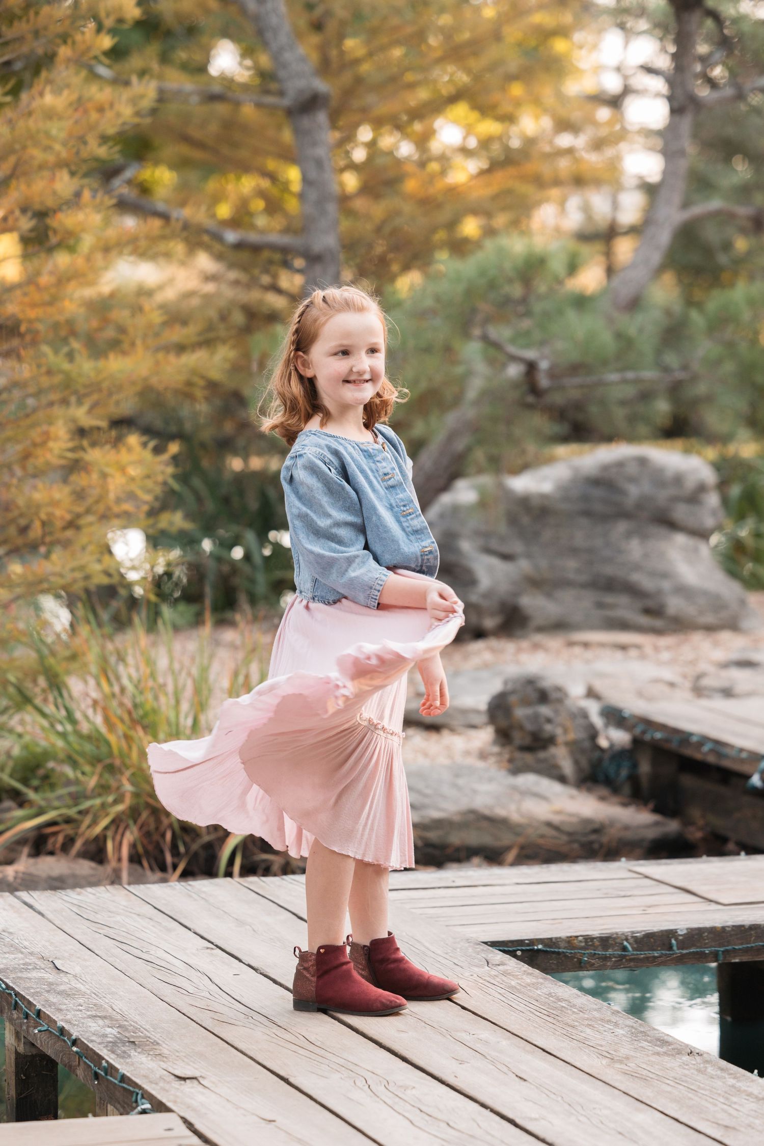 A child in a blue sweater and pink skirt twirls on a wooden dock over water.