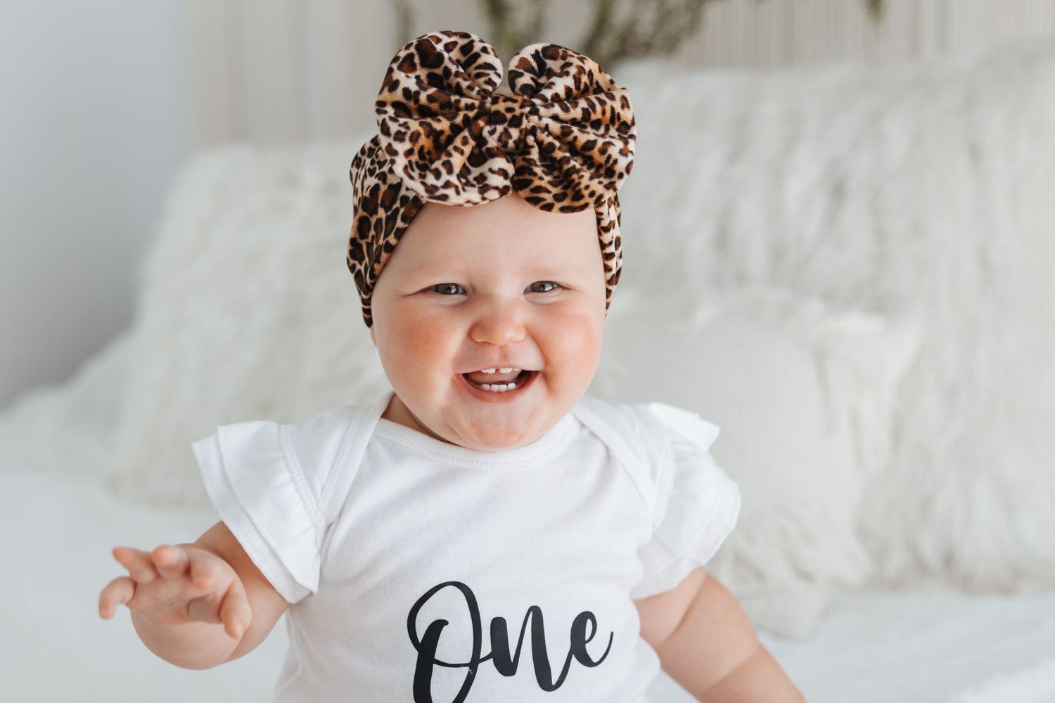 A happy baby in a leopard print bow and white 'One' onesie poses on white bedding.