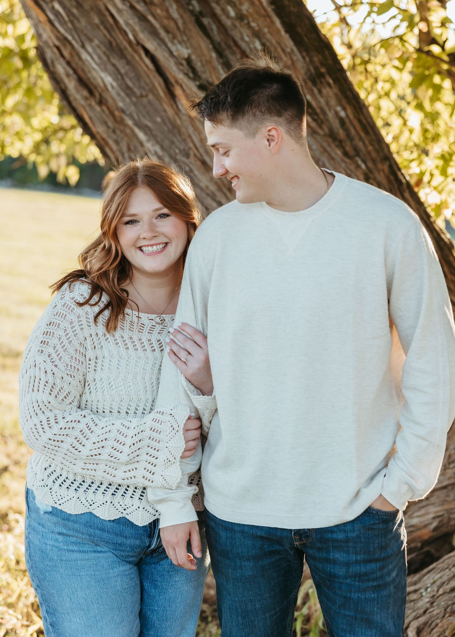 A couple laughing together while standing against a large tree trunk during sunset.
