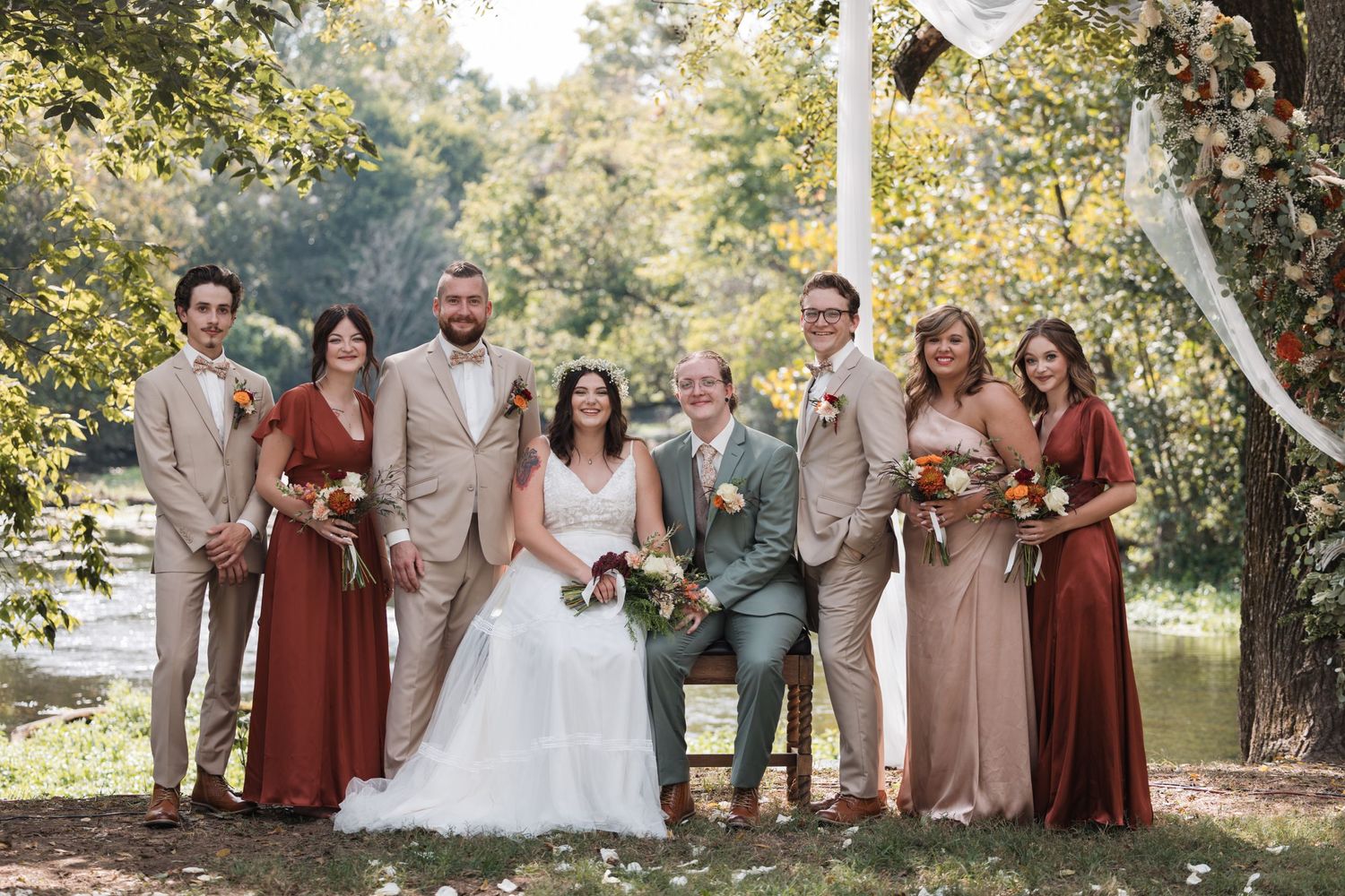 Wedding party in earth-toned dresses and suits poses together under trees.