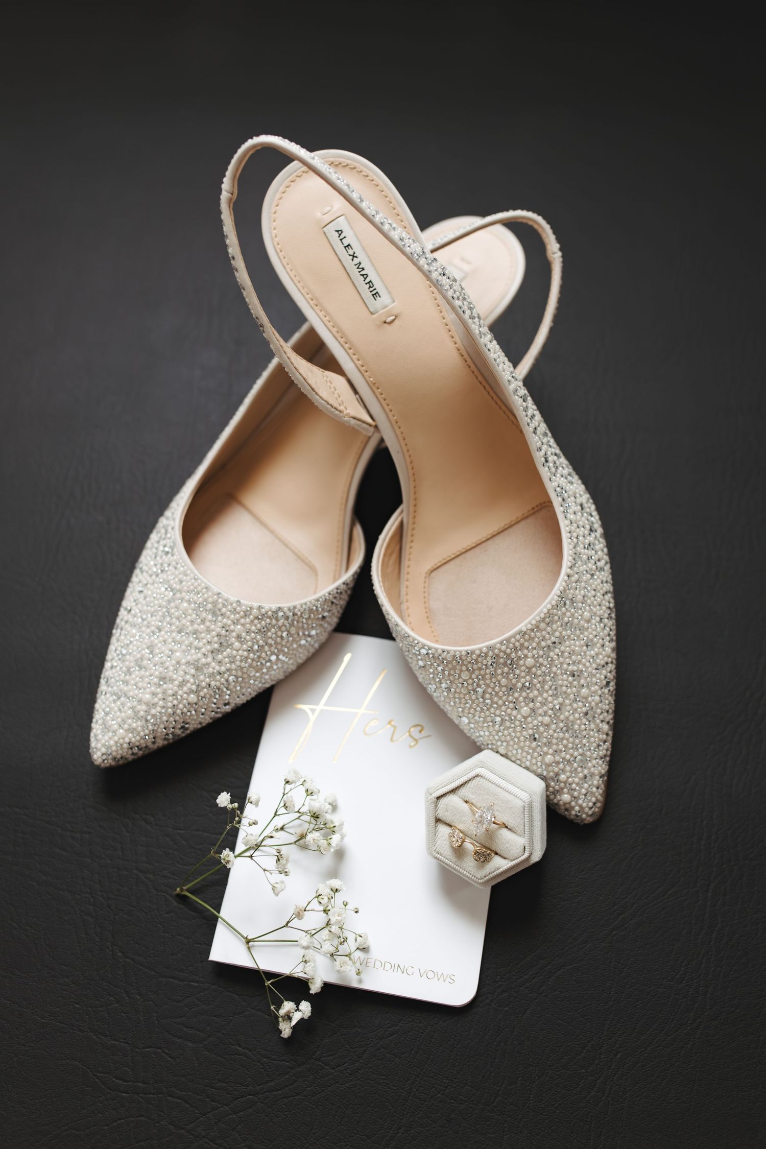 Glittery silver pointed-toe slingback heels with delicate white flowers and a hexagonal ring on dark background.