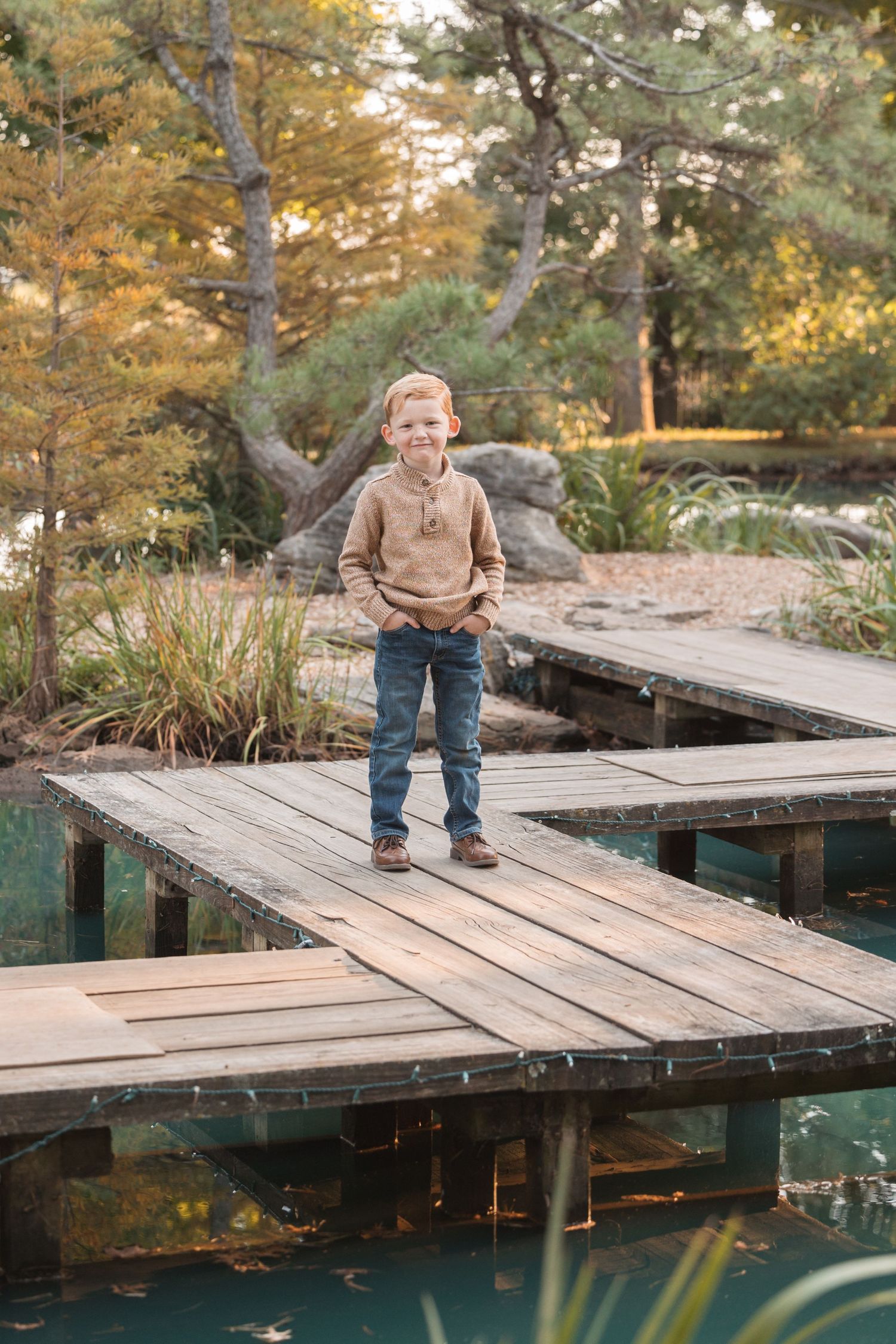 Child in brown sweater stands on wooden dock over tranquil pond surrounded by fall foliage.