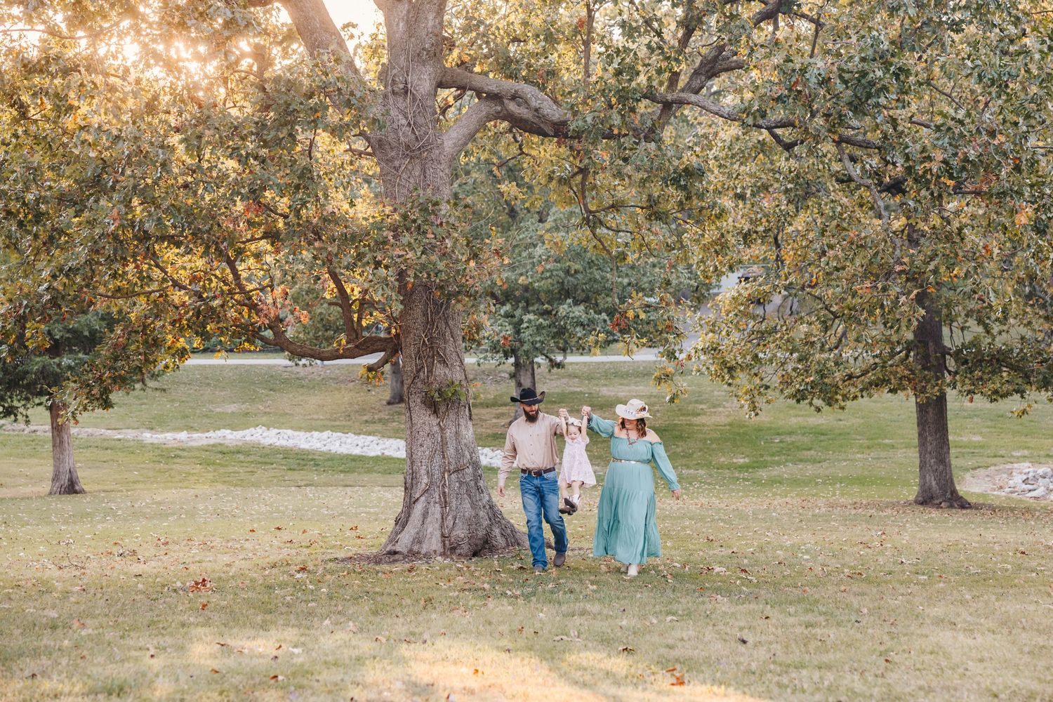 Three figures hold hands while running through a golden sunlit park with mature trees.