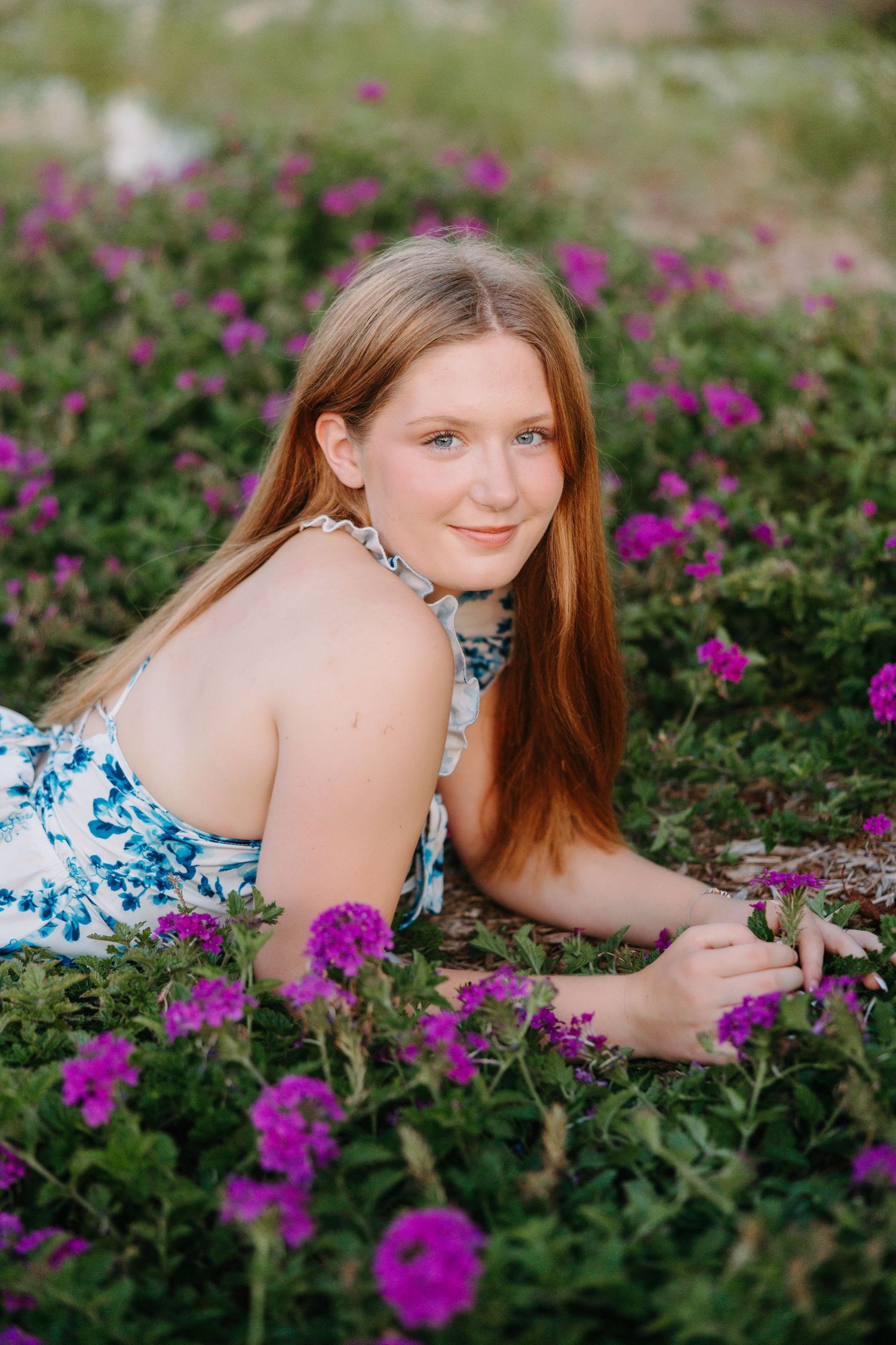 A figure lying in a garden of vibrant purple flowers wearing a white and blue floral dress on a sunny day.