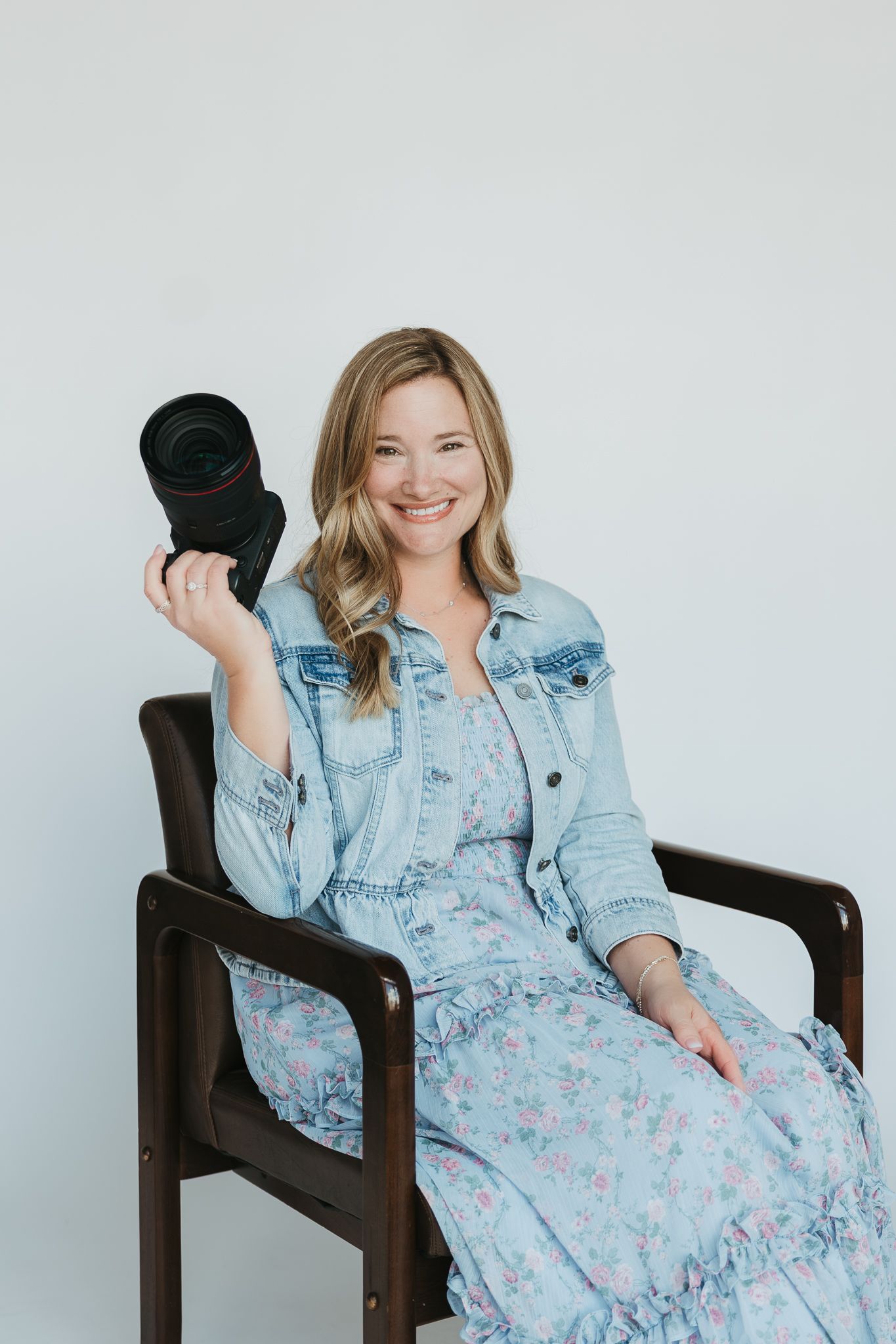 Portrait of photographer in casual denim and floral dress holding black camera lens in studio setting.