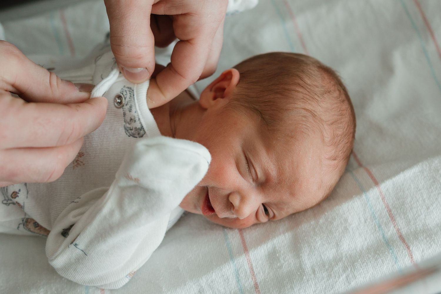 A newborn baby wearing a white outfit lies on white bedding while hands adjust their clothing.