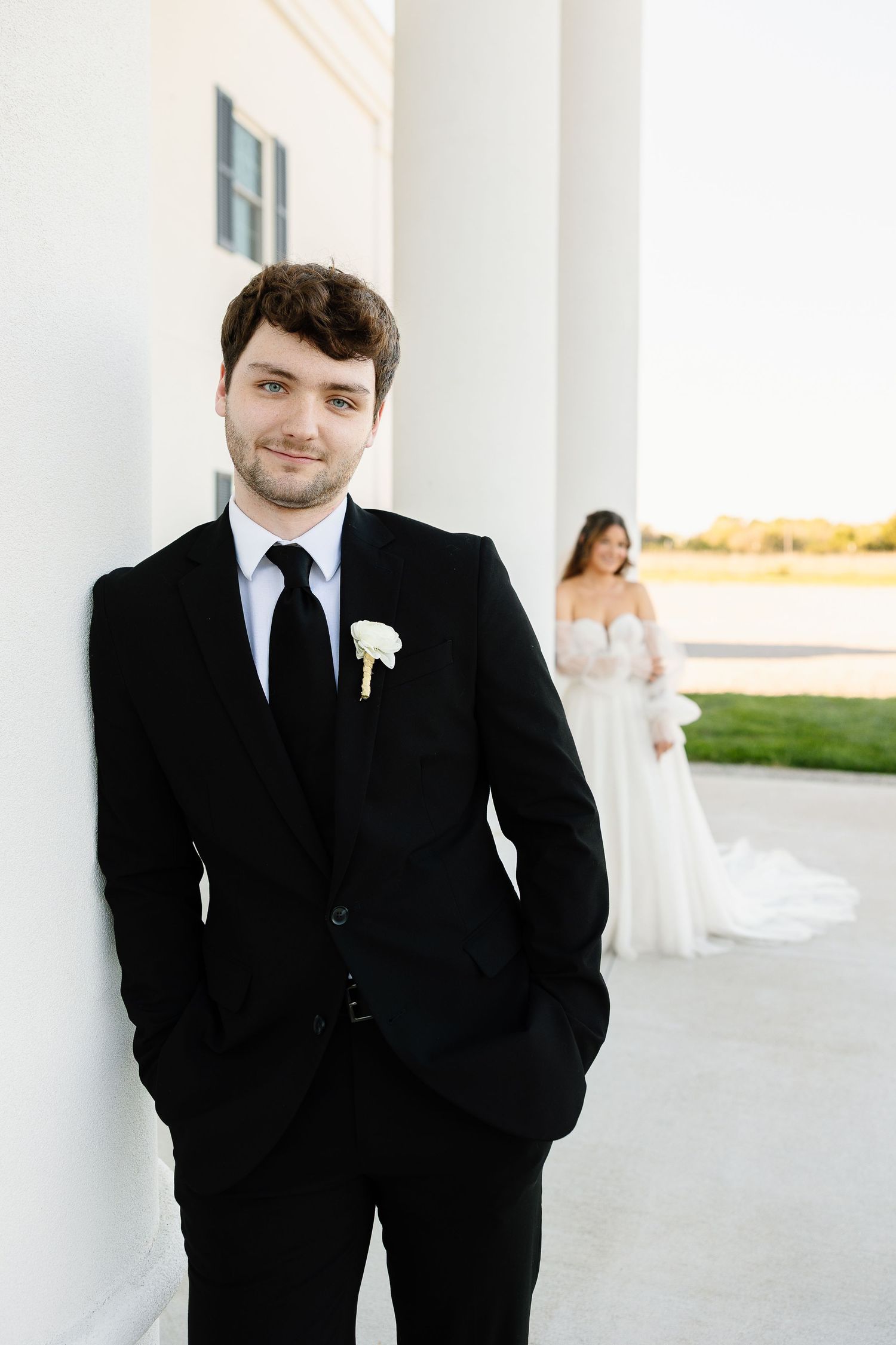 Wedding couple photographed outside white columned building with groom in black suit and bride in white dress in background.