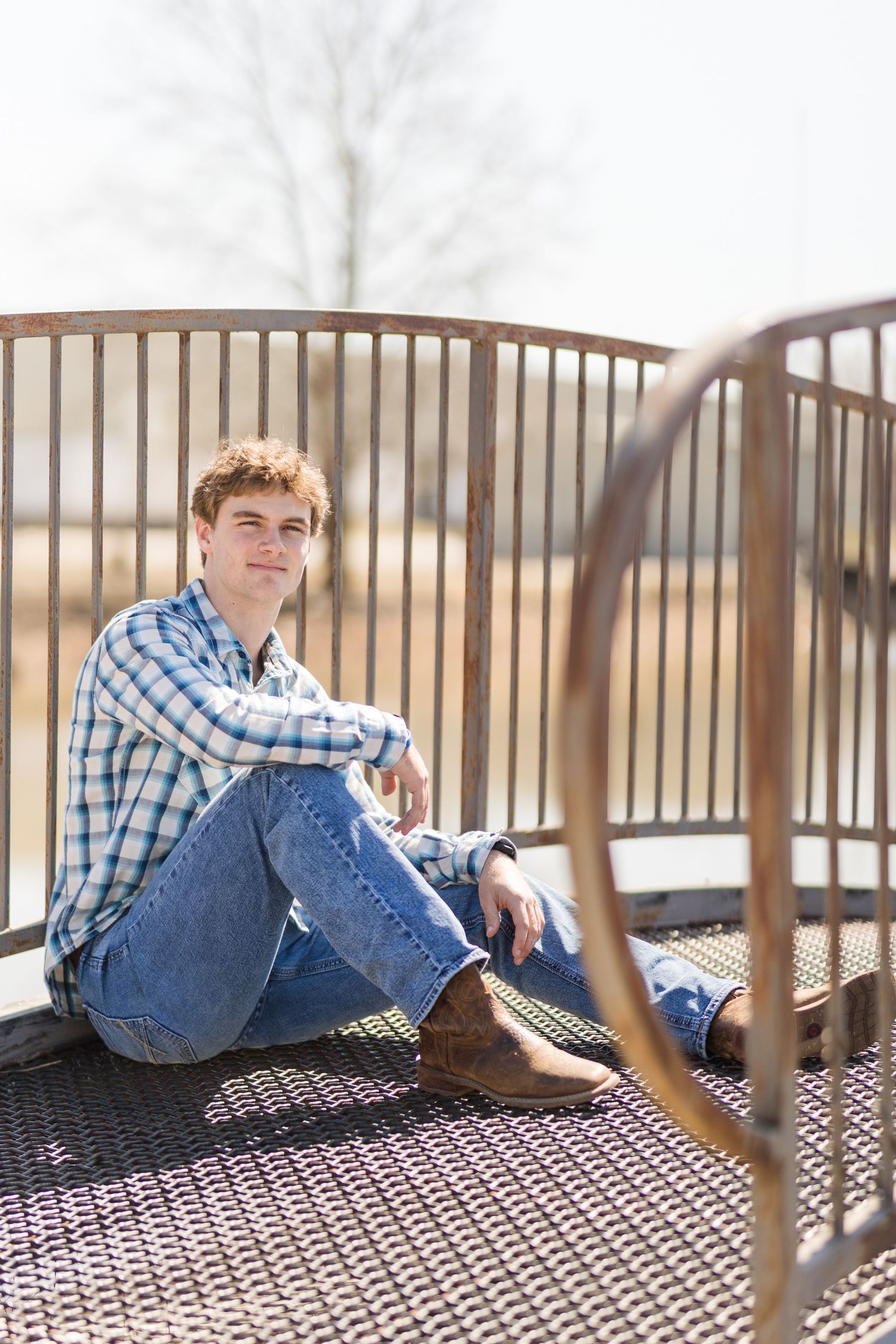 Casual portrait of a person in plaid shirt and jeans sitting on metal platform outdoors.