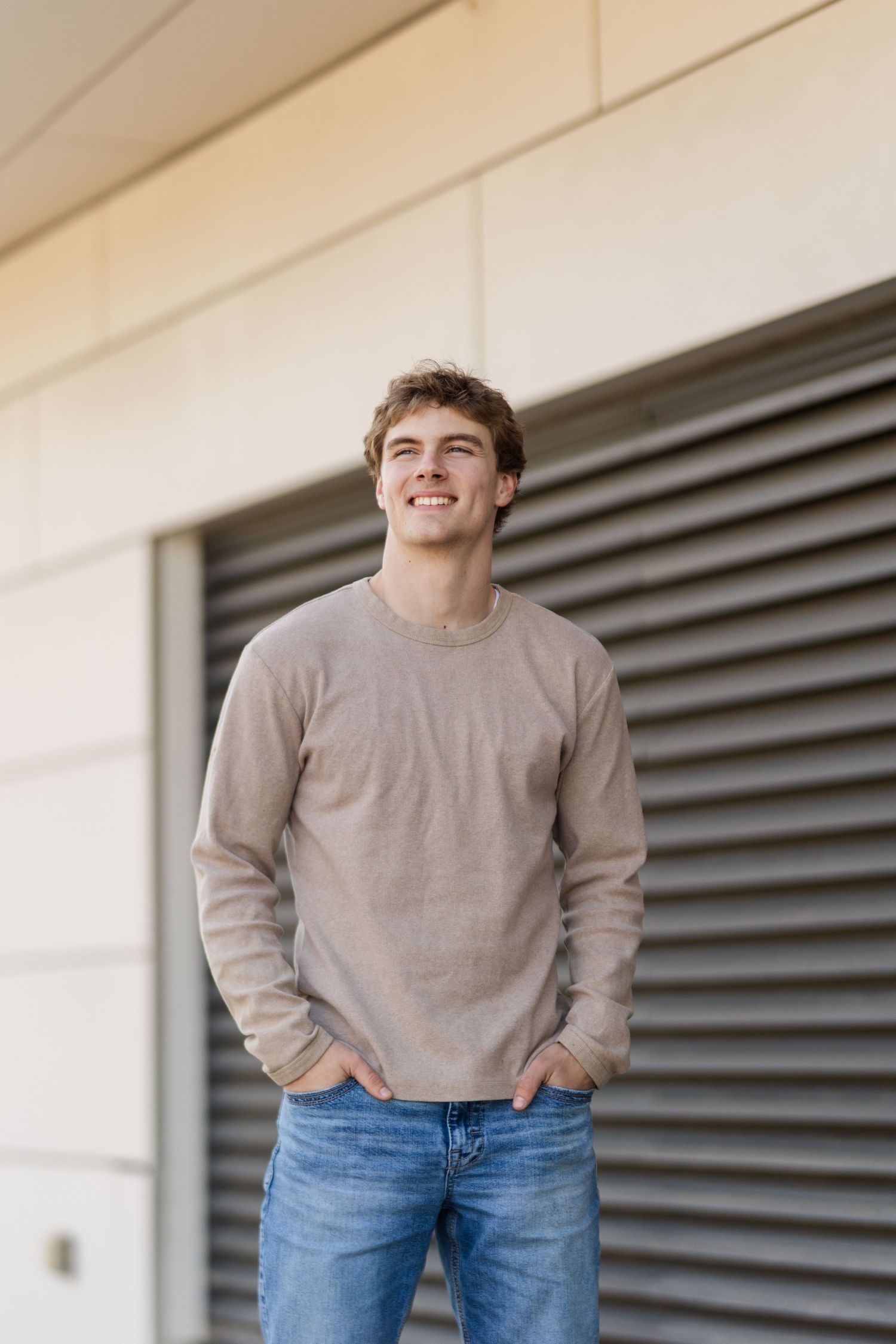 A young person in a tan sweater and blue jeans stands smiling with hands in pockets against a light wall.