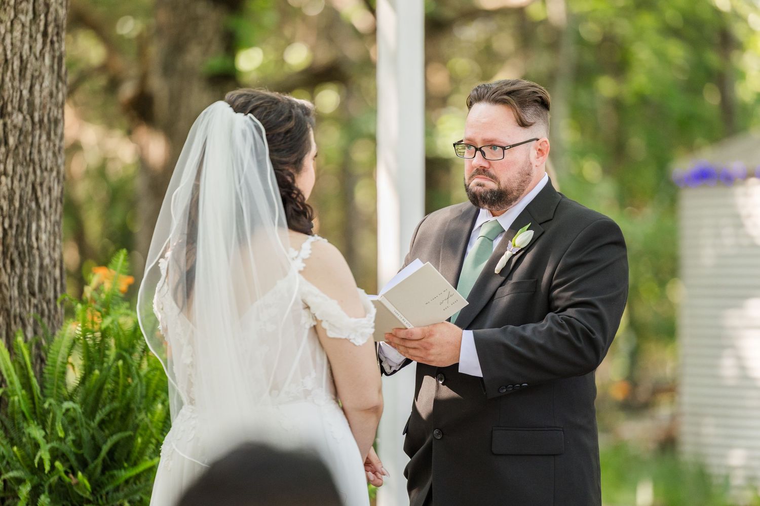Groom reads wedding vows to bride in white dress and veil during outdoor ceremony surrounded by trees.