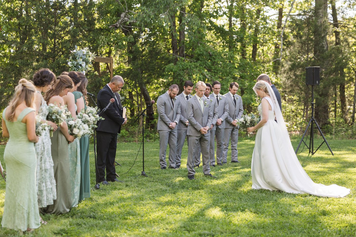 Outdoor wedding ceremony in a lush green forest setting with bridesmaids in sage green dresses and groomsmen in gray suits.