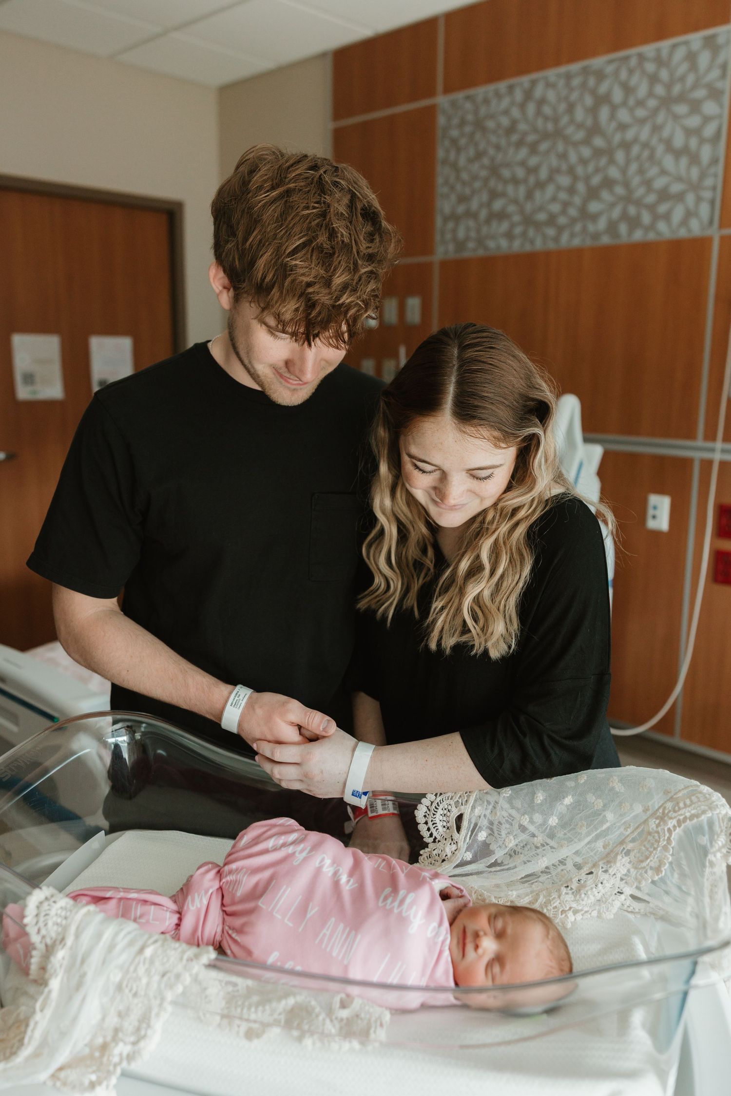 Two people in black shirts look down at a newborn baby wearing pink in a hospital bassinet.