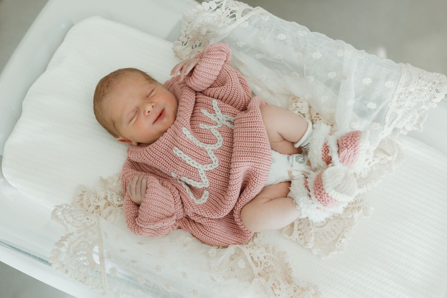 Sleeping newborn baby wearing soft pink knit sweater and booties lying on white lace blanket.