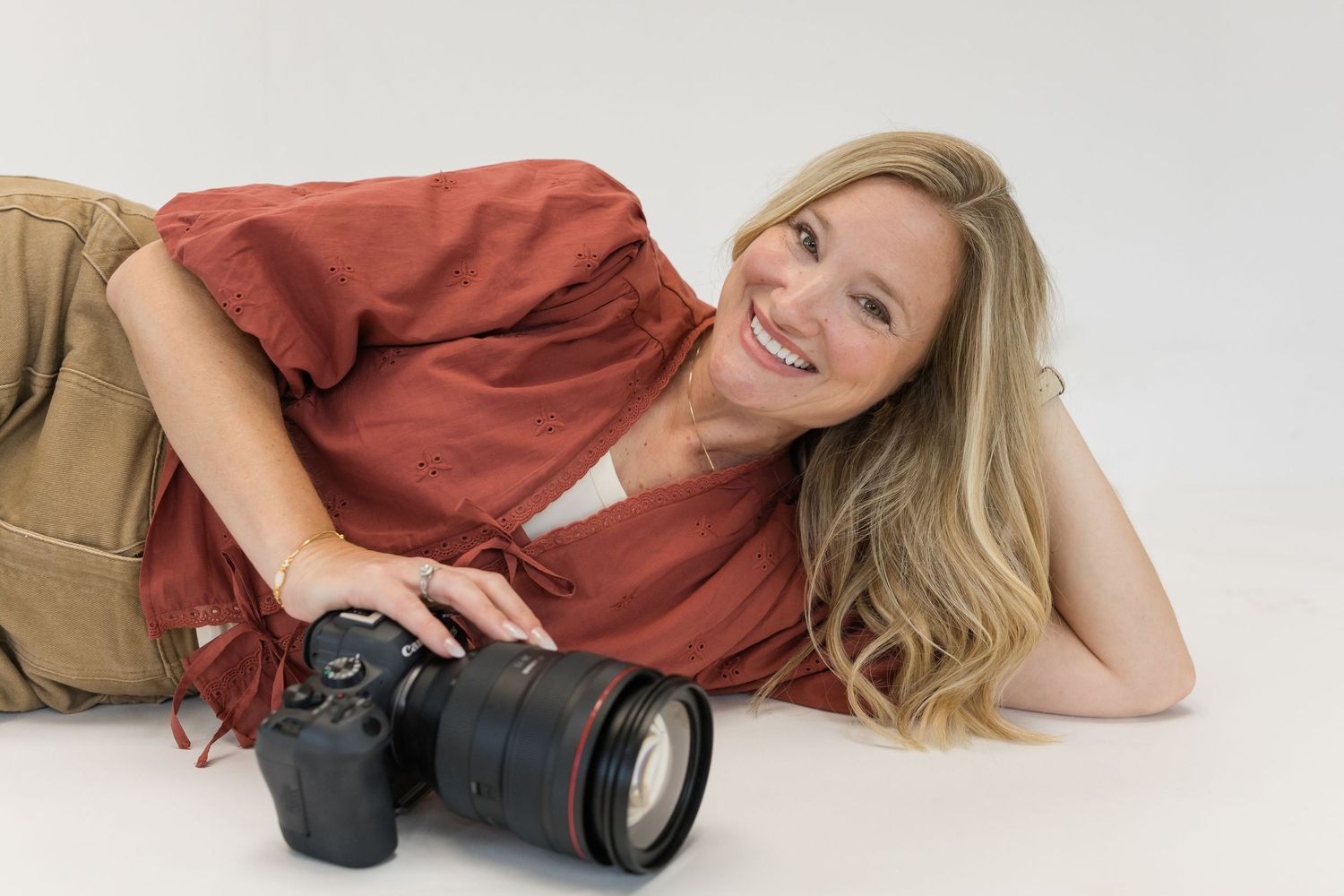 Smiling blonde woman lying down holding a Canon DSLR camera with zoom lens on white background.