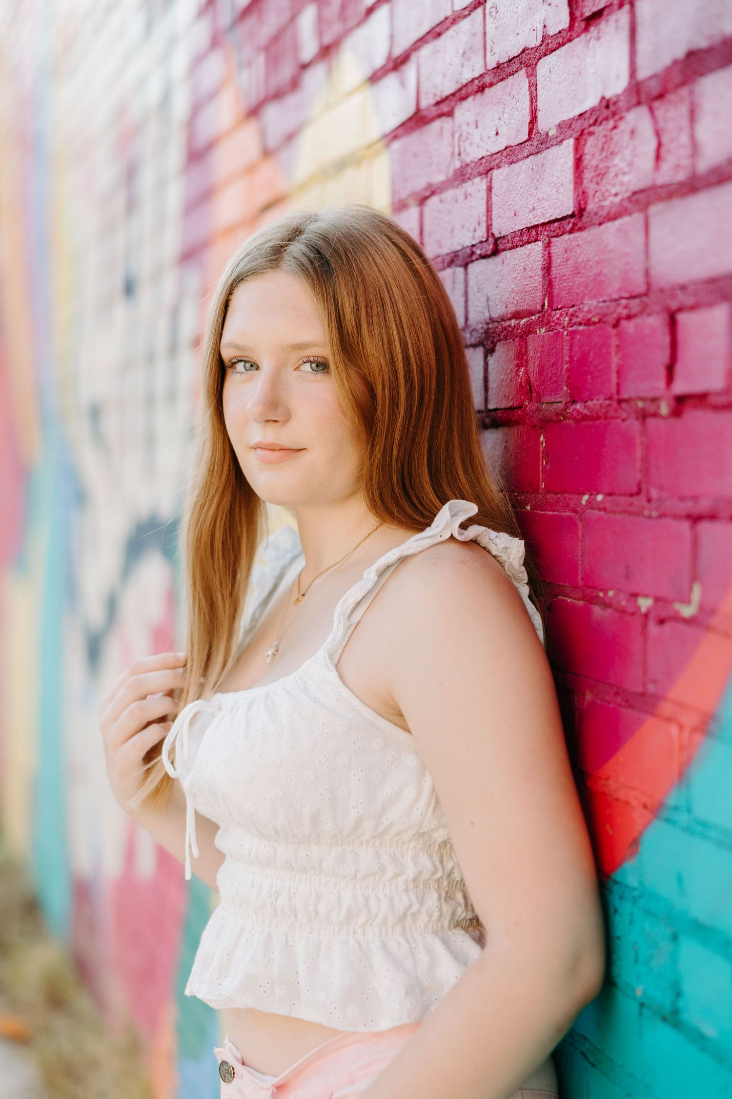 A model in a white ruffled top poses artistically against a bright pink and turquoise painted brick wall.