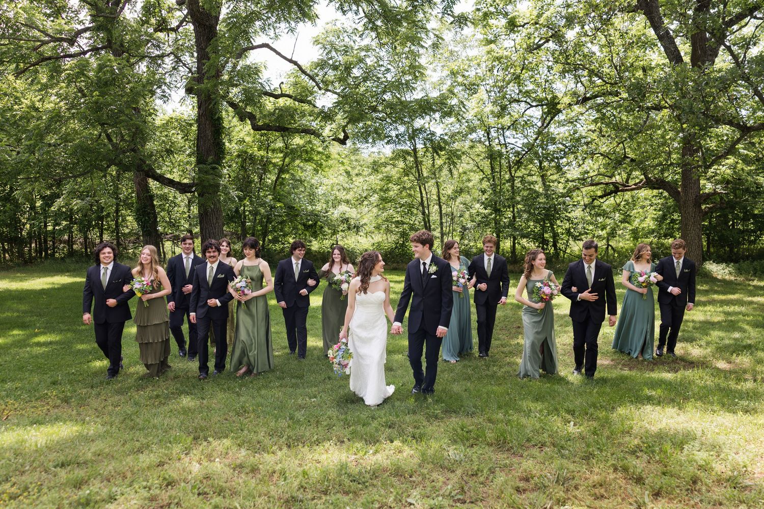 Wedding party walking together outdoors in a lush green forest, bridesmaids in sage green dresses.
