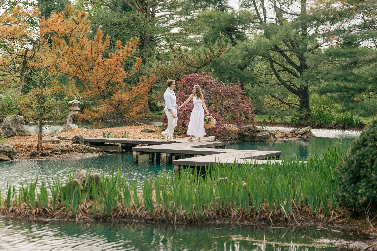 Couple holding hands on a wooden dock bridge over a serene pond in a lush Japanese garden during autumn.