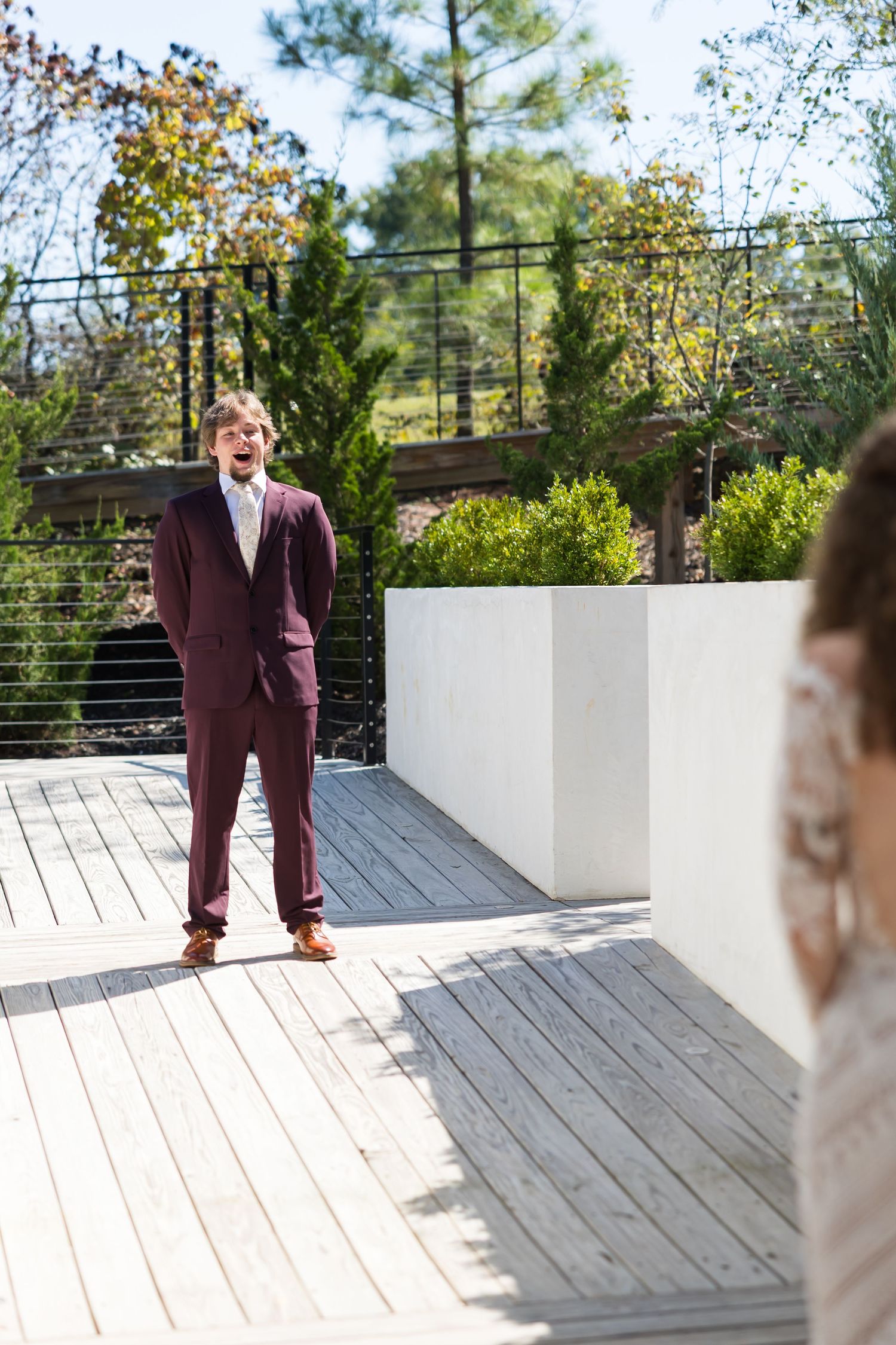 Groom in burgundy suit reacts emotionally during first look with bride on outdoor wooden deck.