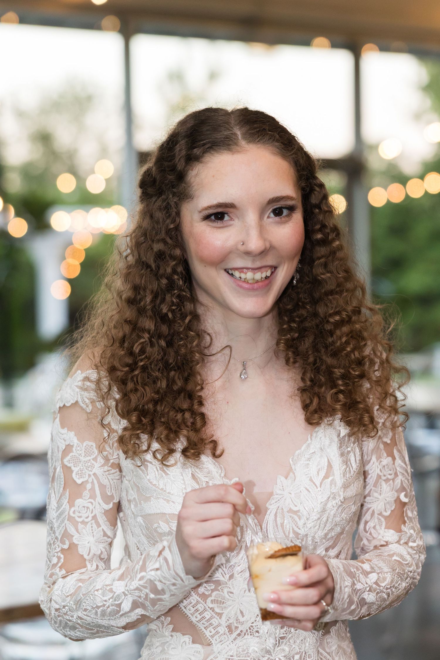 Smiling curly-haired bride in lace wedding dress holds a small cup at a reception with string lights.