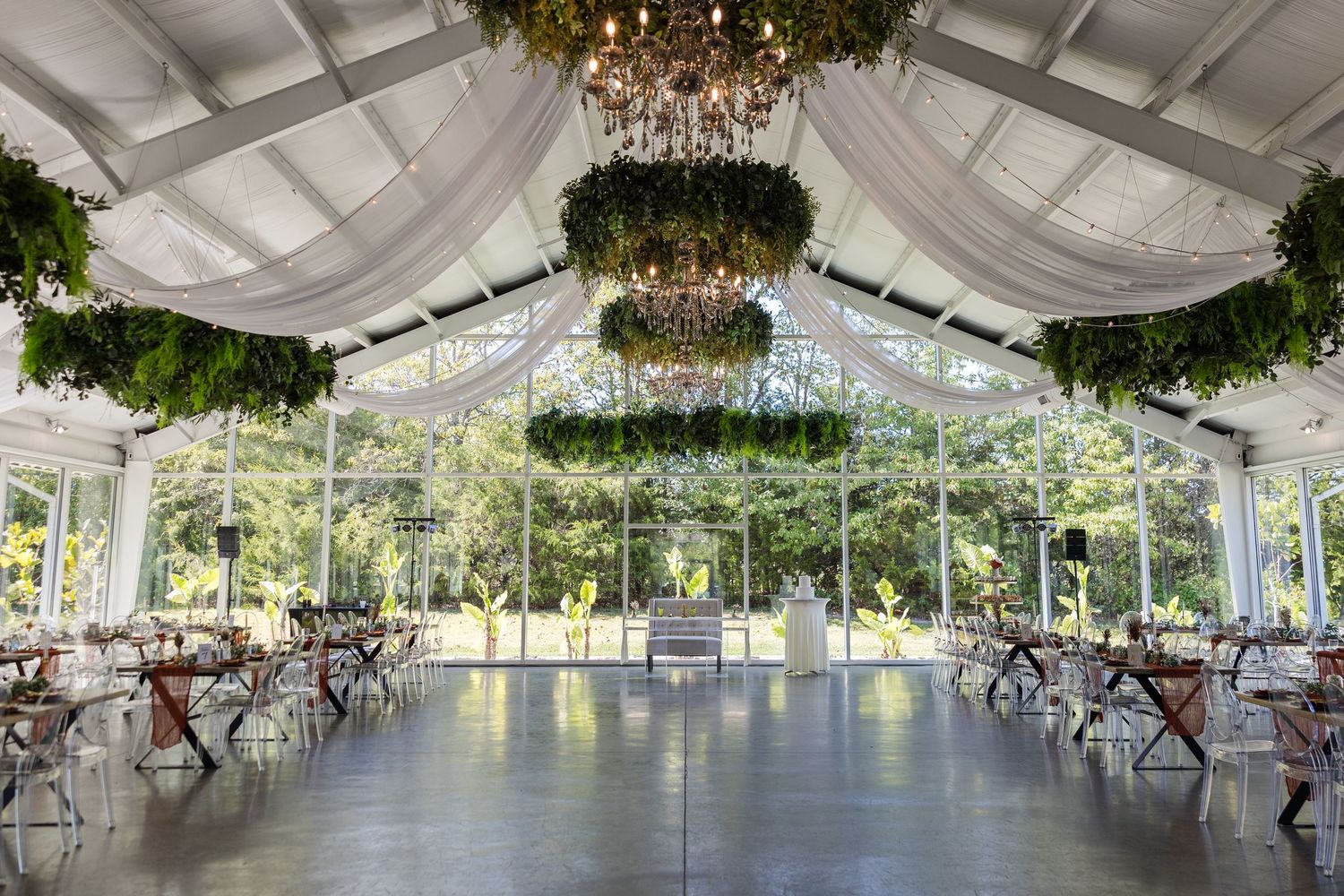 Elegant greenhouse wedding venue with chandeliers, lush greenery, white draping, and floor-to-ceiling windows.