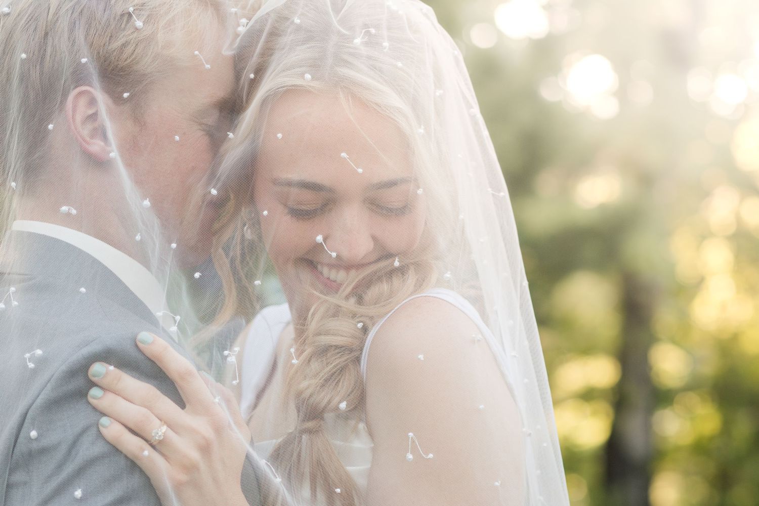 A bride and groom share a tender moment under a flowing veil, smiling softly in warm bokeh outdoor light.